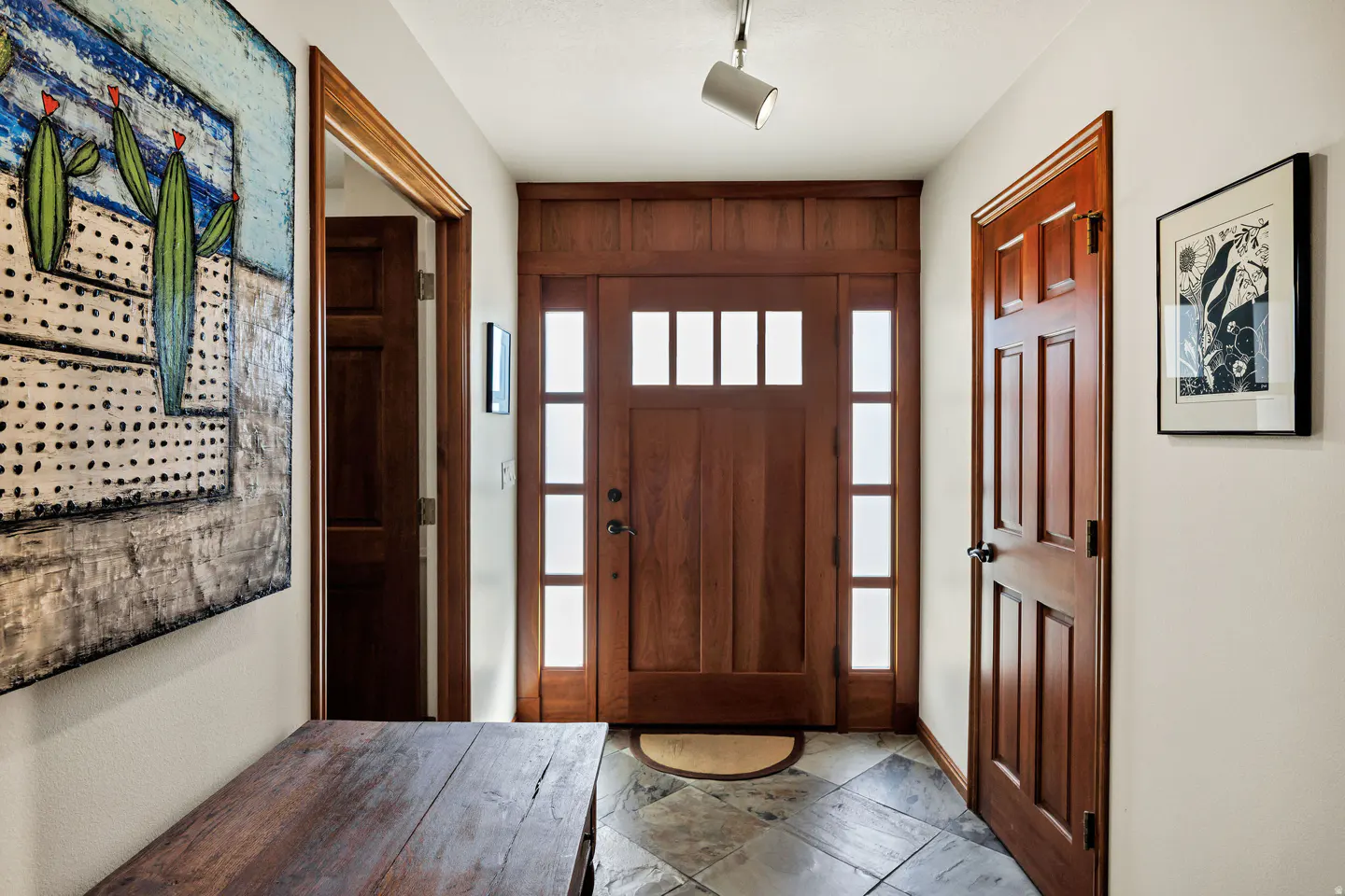Entryway with a wooden front door, sidelights, and transom windows. A cactus painting hangs above a wooden bench. Slate flooring and white walls.