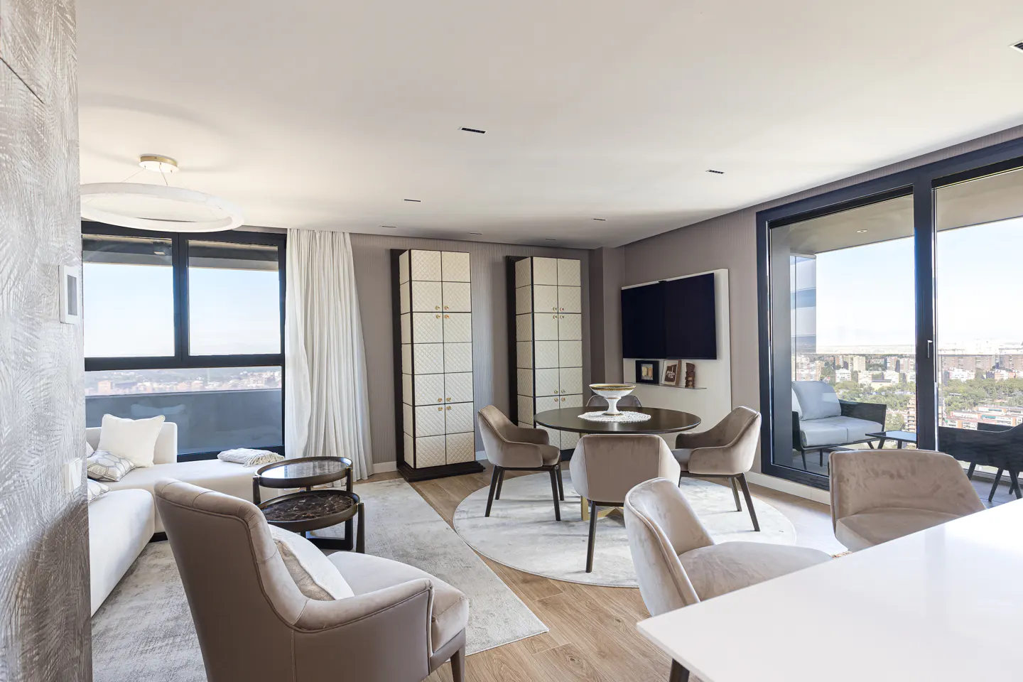 A modern living room with a round dining table, beige chairs, and a balcony with city views. Two tall cabinets stand against a gray wall.