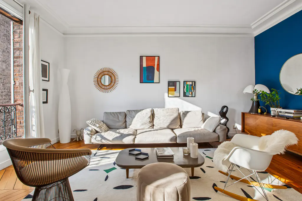 Bright living room with a gray sofa, modern art, and a white rocking chair on a patterned rug. A blue accent wall adds a pop of color.