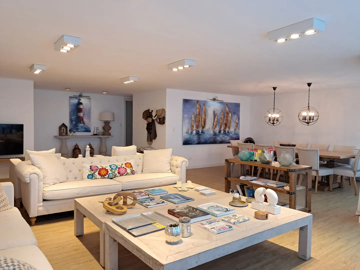 Bright, open living room with white sofas, a large coffee table with books, and nautical-themed art. Dining area visible in the background.