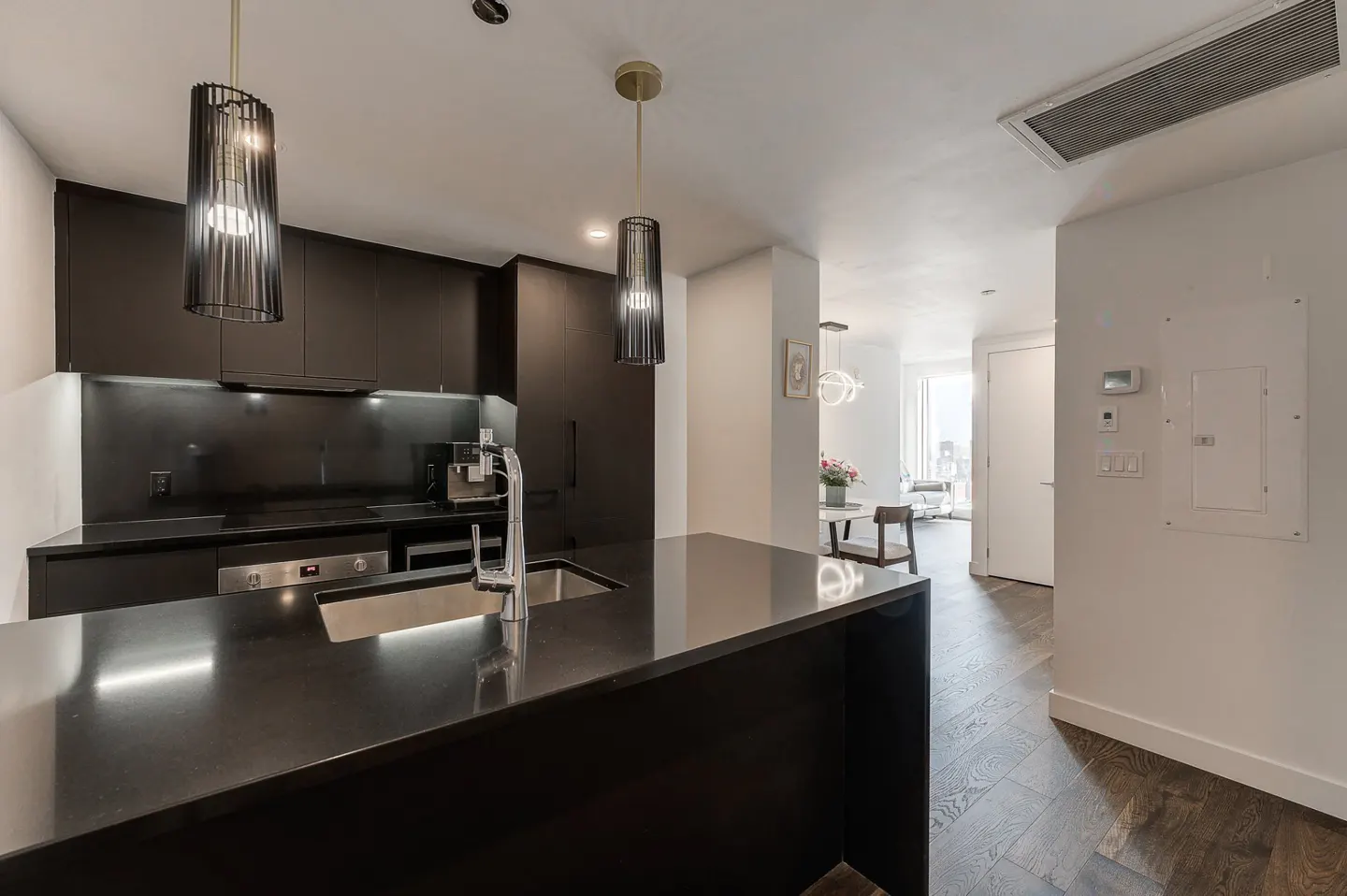 Modern kitchen with black cabinets, island, and pendant lights. Dining area visible in the background with white walls and wood floors.