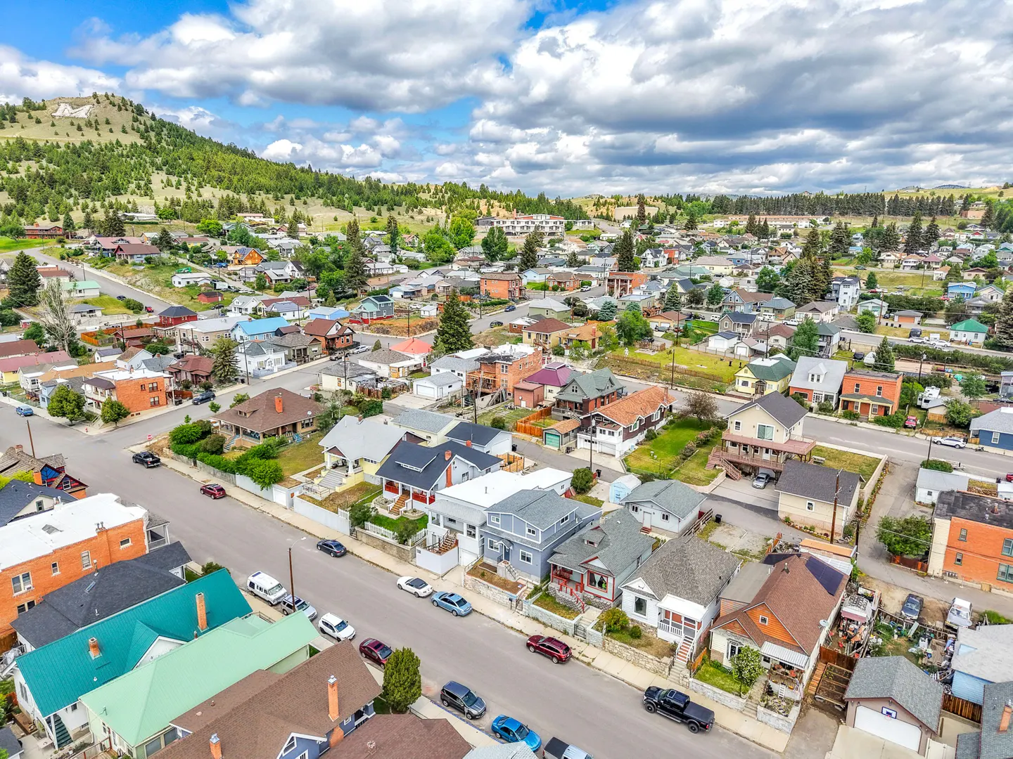 Aerial view of a residential neighborhood with colorful houses, cars on the street, and a green hill in the background under a cloudy sky.