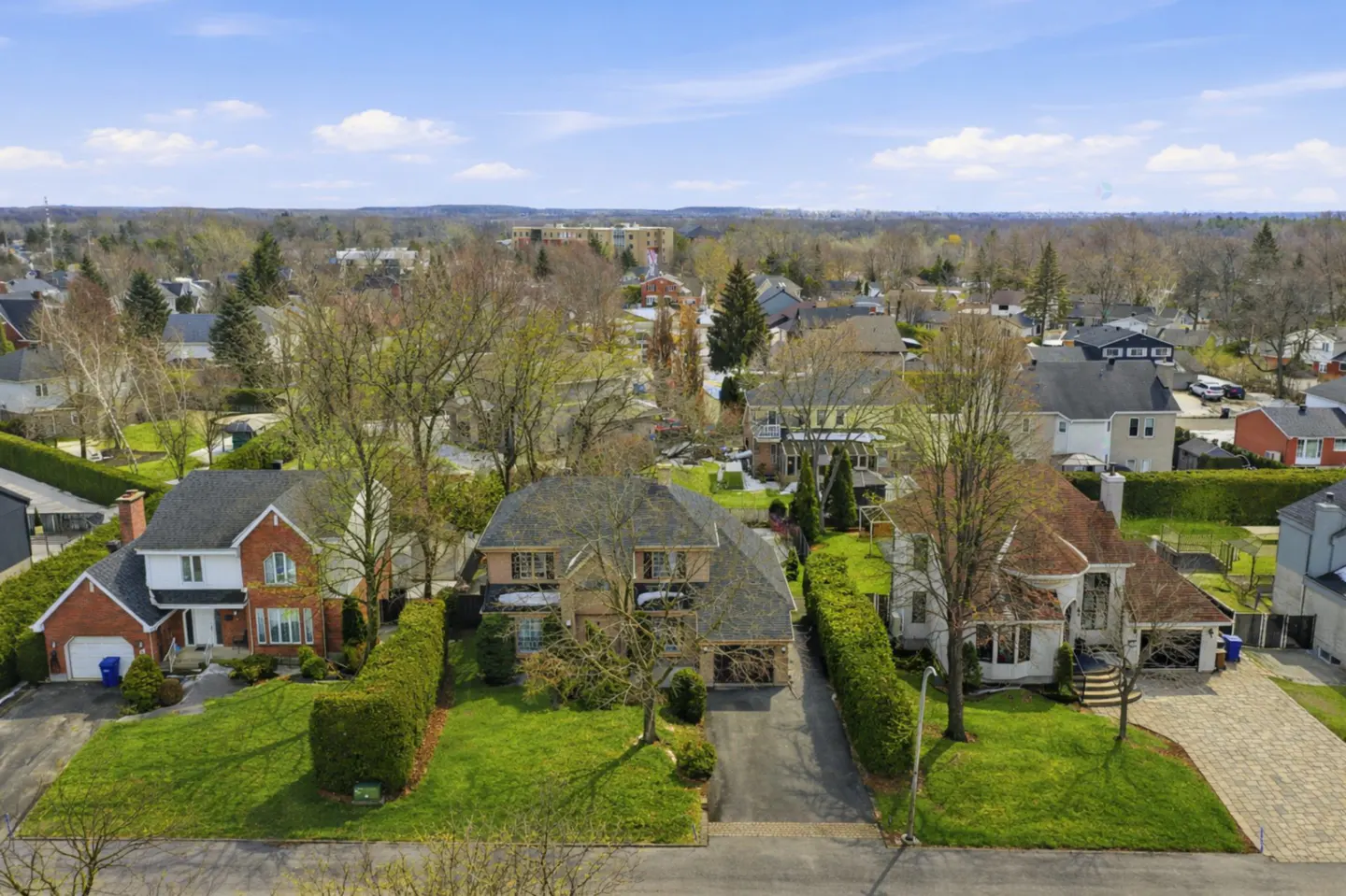 Aerial view of a suburban neighborhood with houses, green lawns, trees, and a blue sky with clouds.