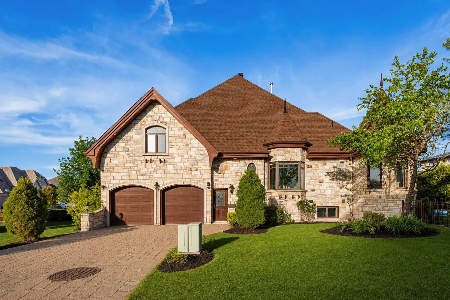 Two-story stone house with brown roof, two-car garage, and green lawn under a blue sky.