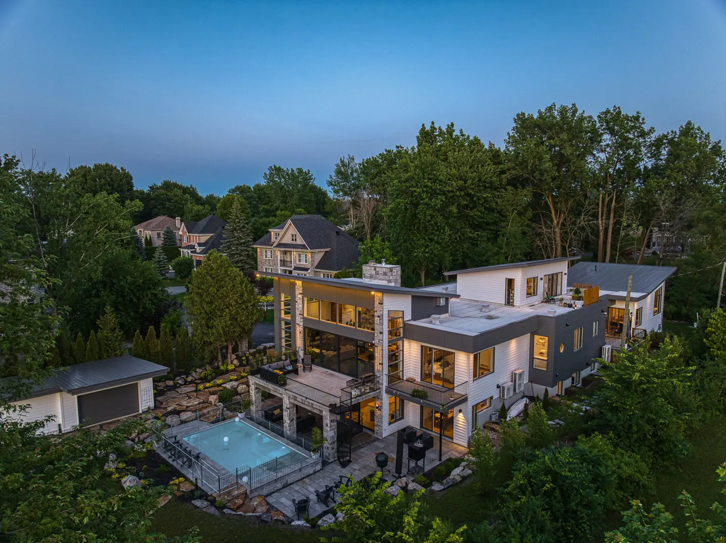Aerial view of a modern, two-story house with a pool, stone patio, and lush green landscaping at dusk.