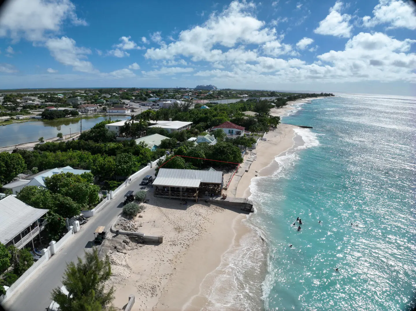 Aerial view of a beachside property with turquoise water, white sand, and a building with a gray roof. People are swimming in the ocean.