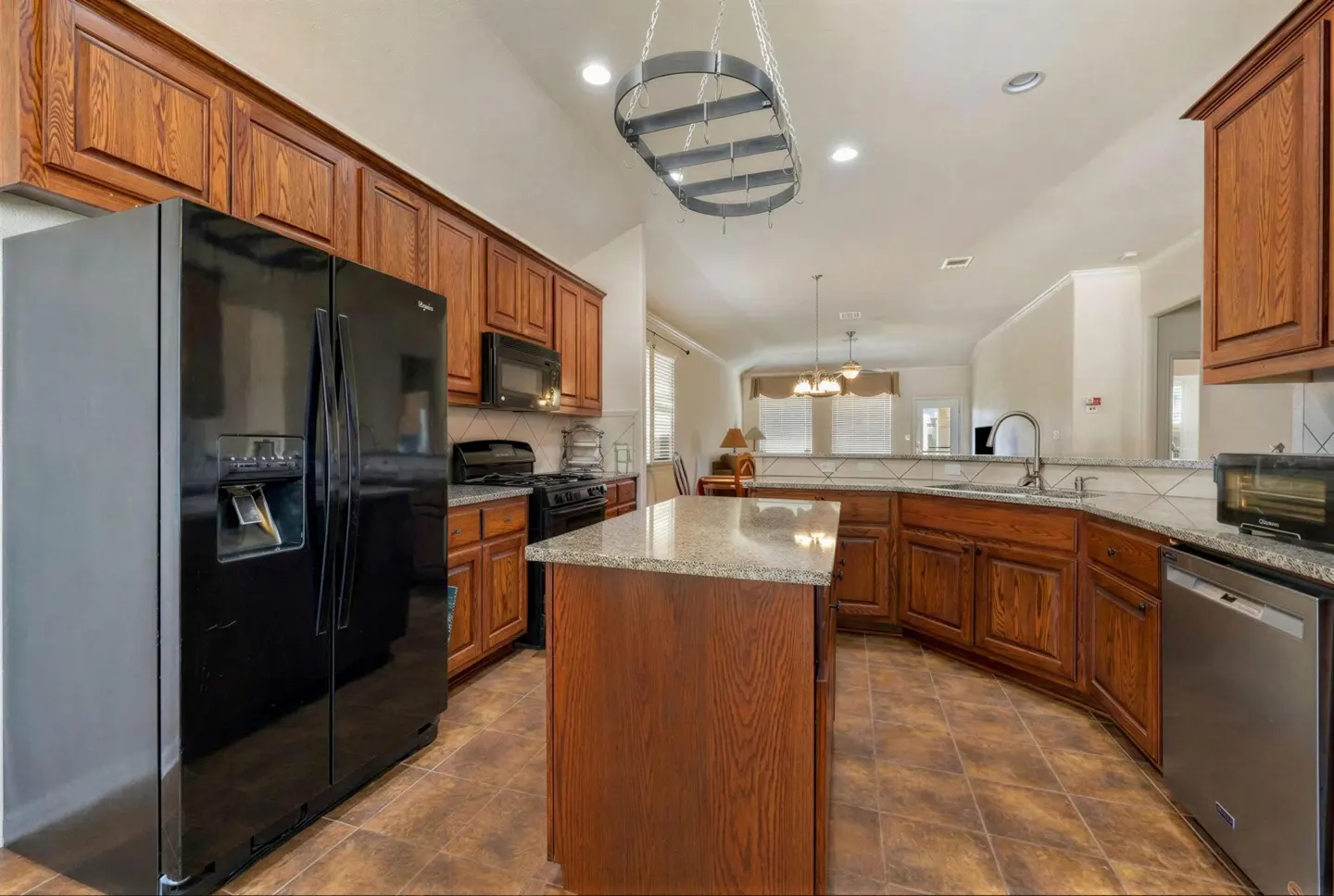 A kitchen with wood cabinets, granite countertops, and stainless steel appliances. A black refrigerator is on the left.