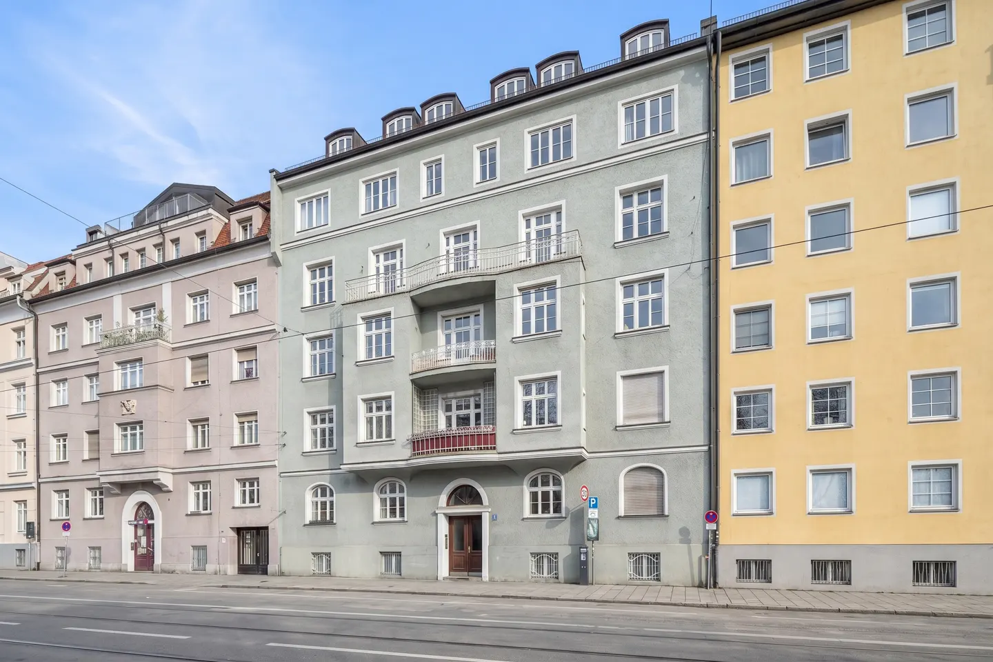 Street view of a row of multi-story buildings in Munich, Germany. The buildings are light yellow, gray, and light pink.