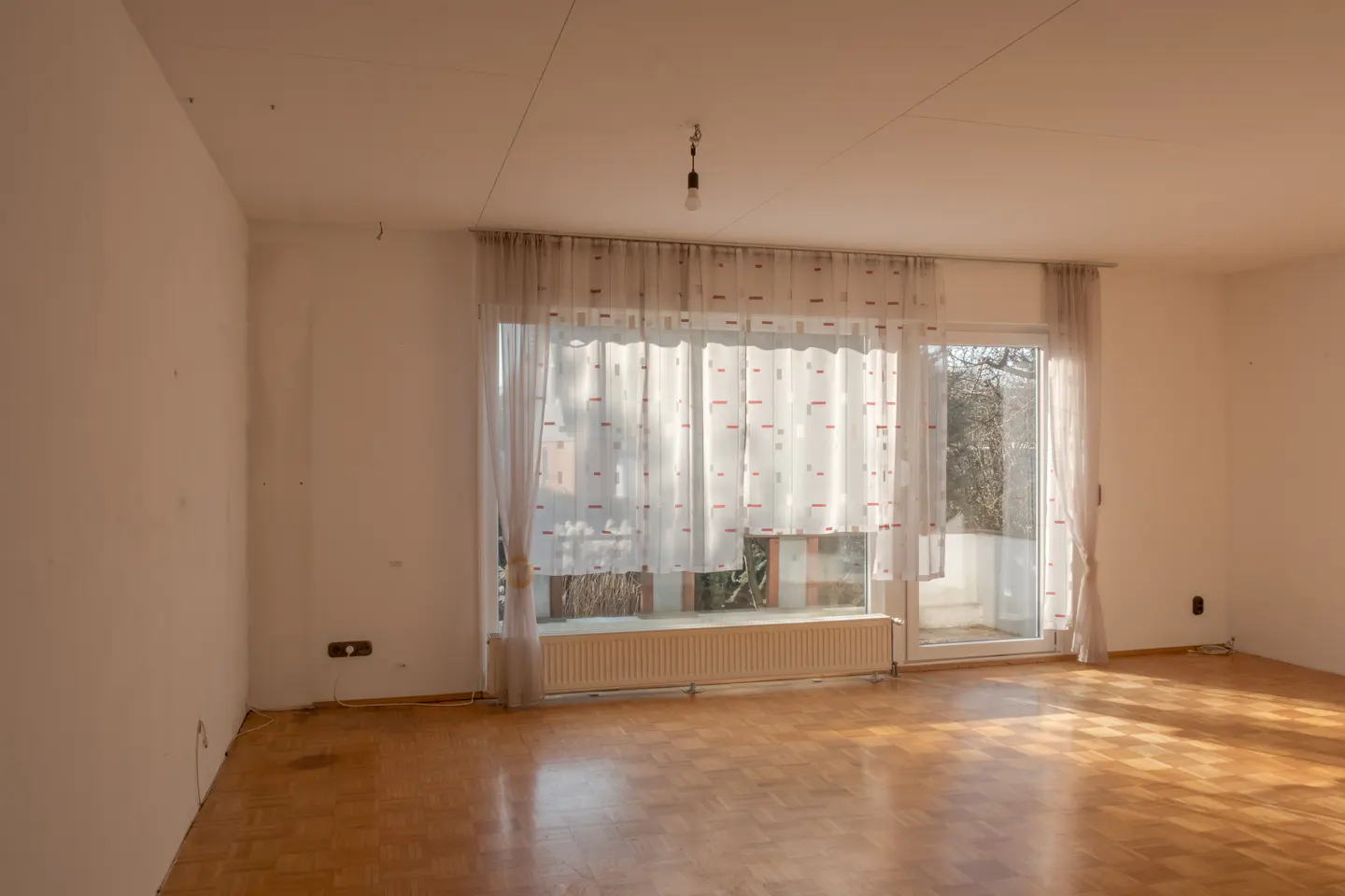 Empty room with parquet floor, white walls, and a large window with sheer curtains. A radiator sits beneath the window. Sunlight streams in.