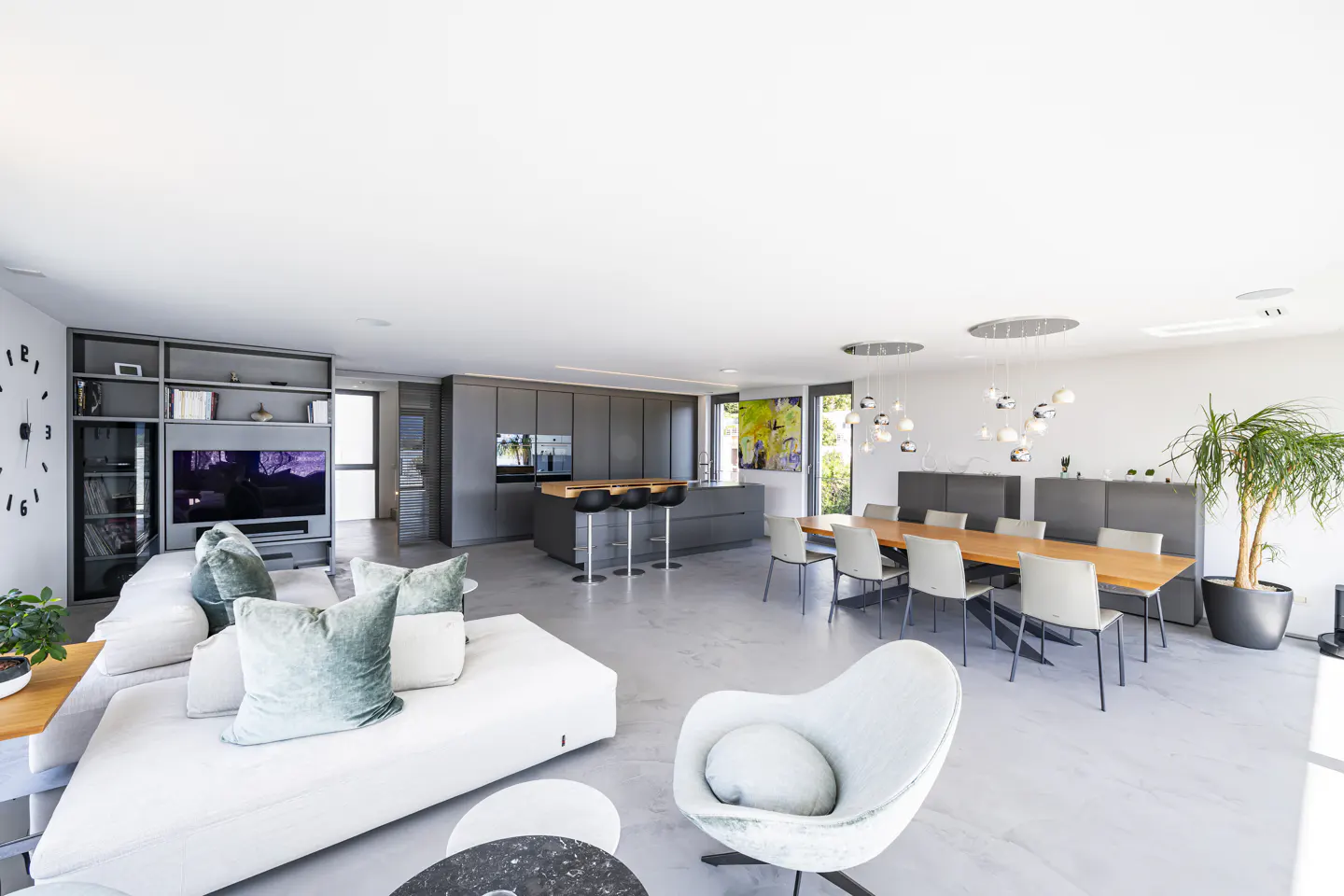 Open-concept living space with white sofa, gray kitchen, wood dining table, and modern lighting. A large clock is on the wall.