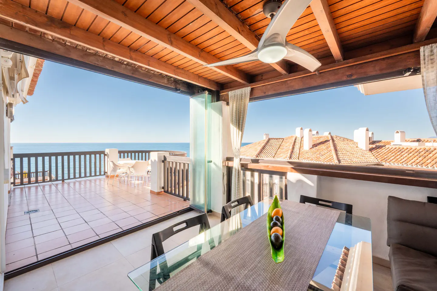 Covered patio with a wood ceiling, ceiling fan, and glass table. Ocean view with a balcony and white chairs in the background.