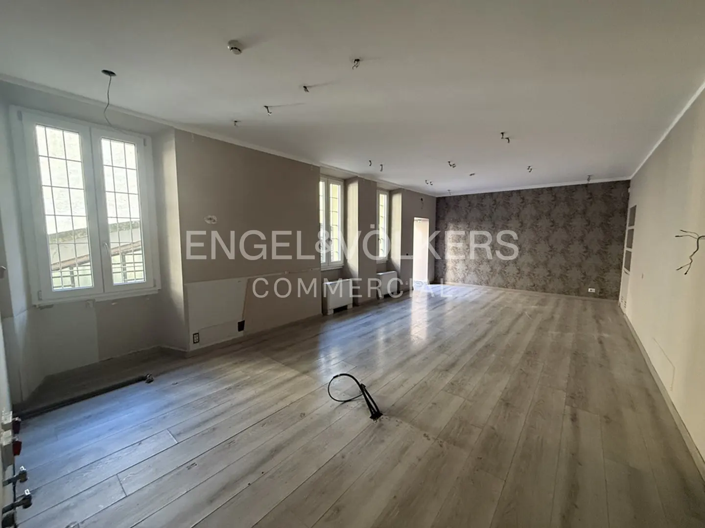 Unfurnished room with light wood floors, beige walls, and large windows. Wallpaper accent wall and Engel & Volkers logo visible.