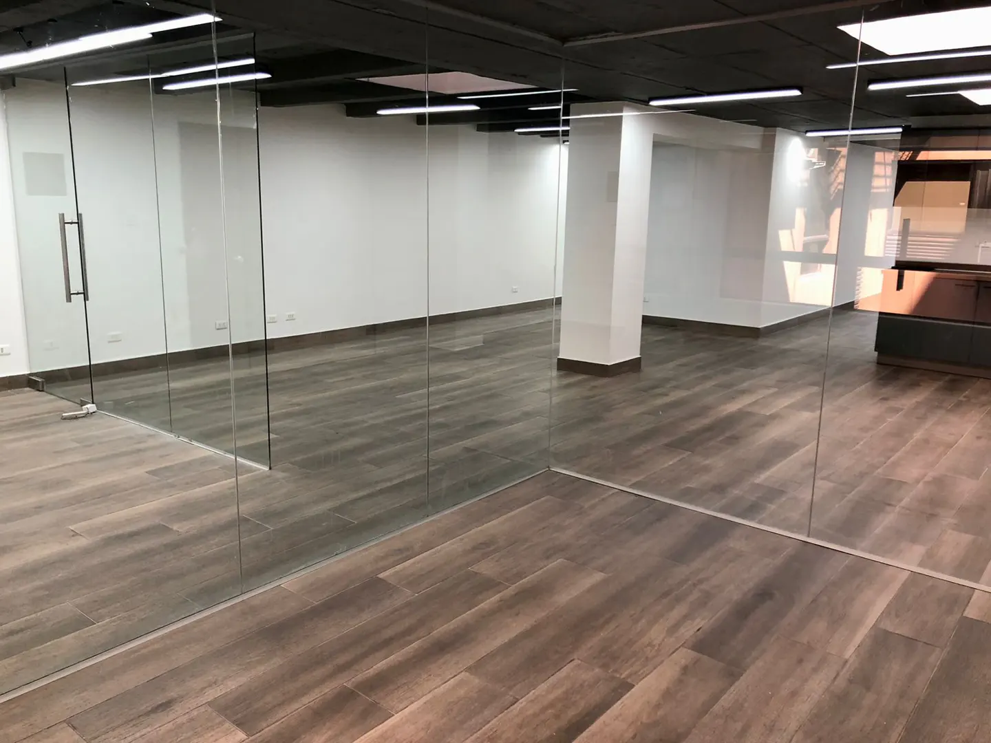 Empty office space with wood-look tile floors, white walls, and a black ceiling. Glass walls and a door enclose the space.