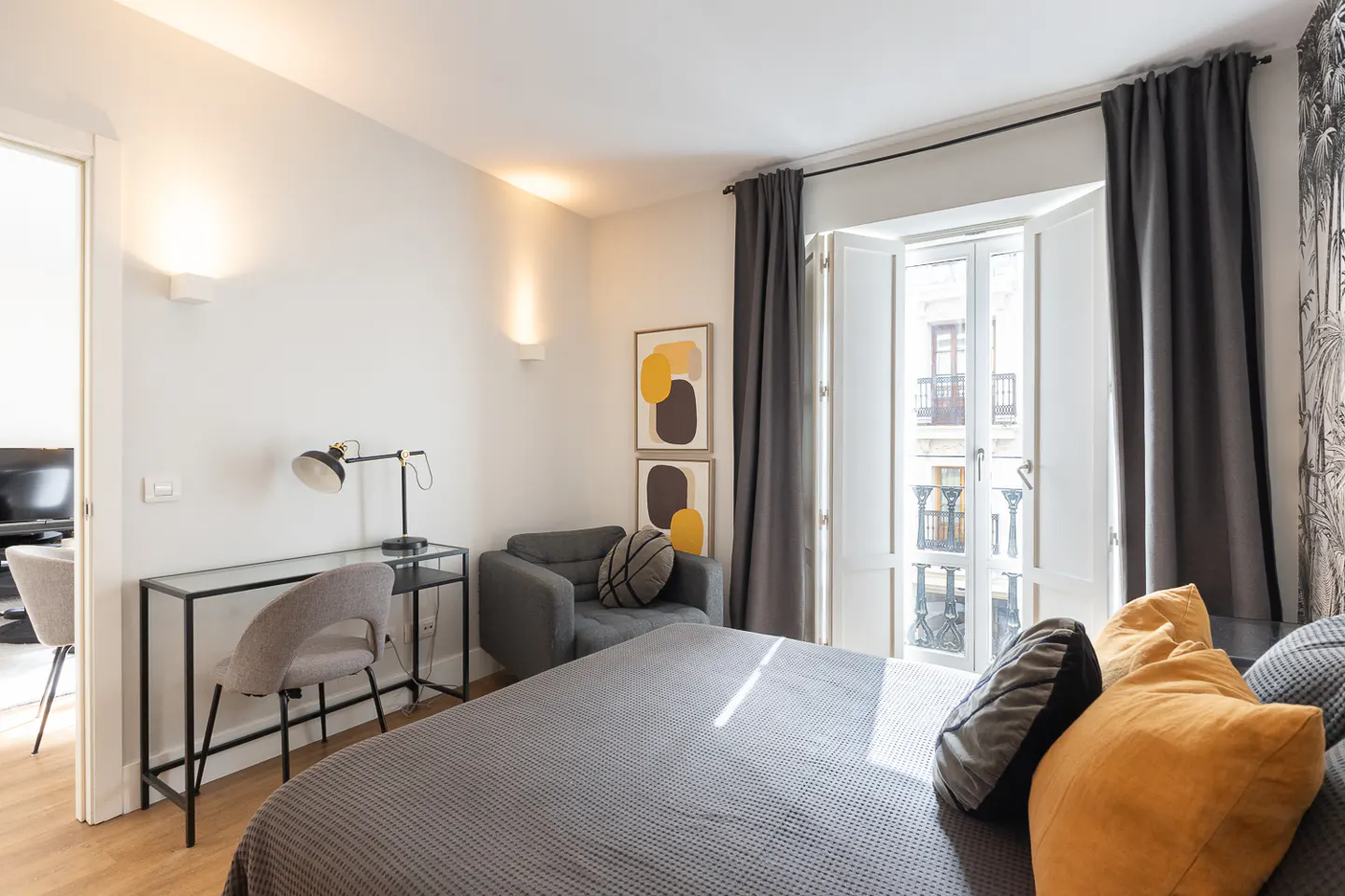 Bedroom with gray bed, yellow pillows, desk, chair, and gray armchair. White walls, abstract art, and open window with gray curtains.
