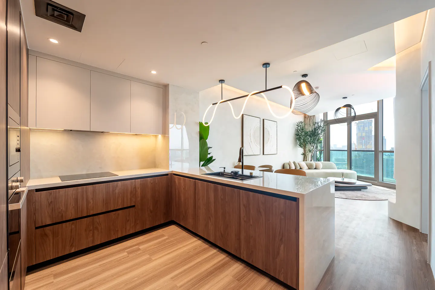 A modern kitchen with wood cabinets, a white countertop, and a black sink. The living room is visible in the background with a large window.