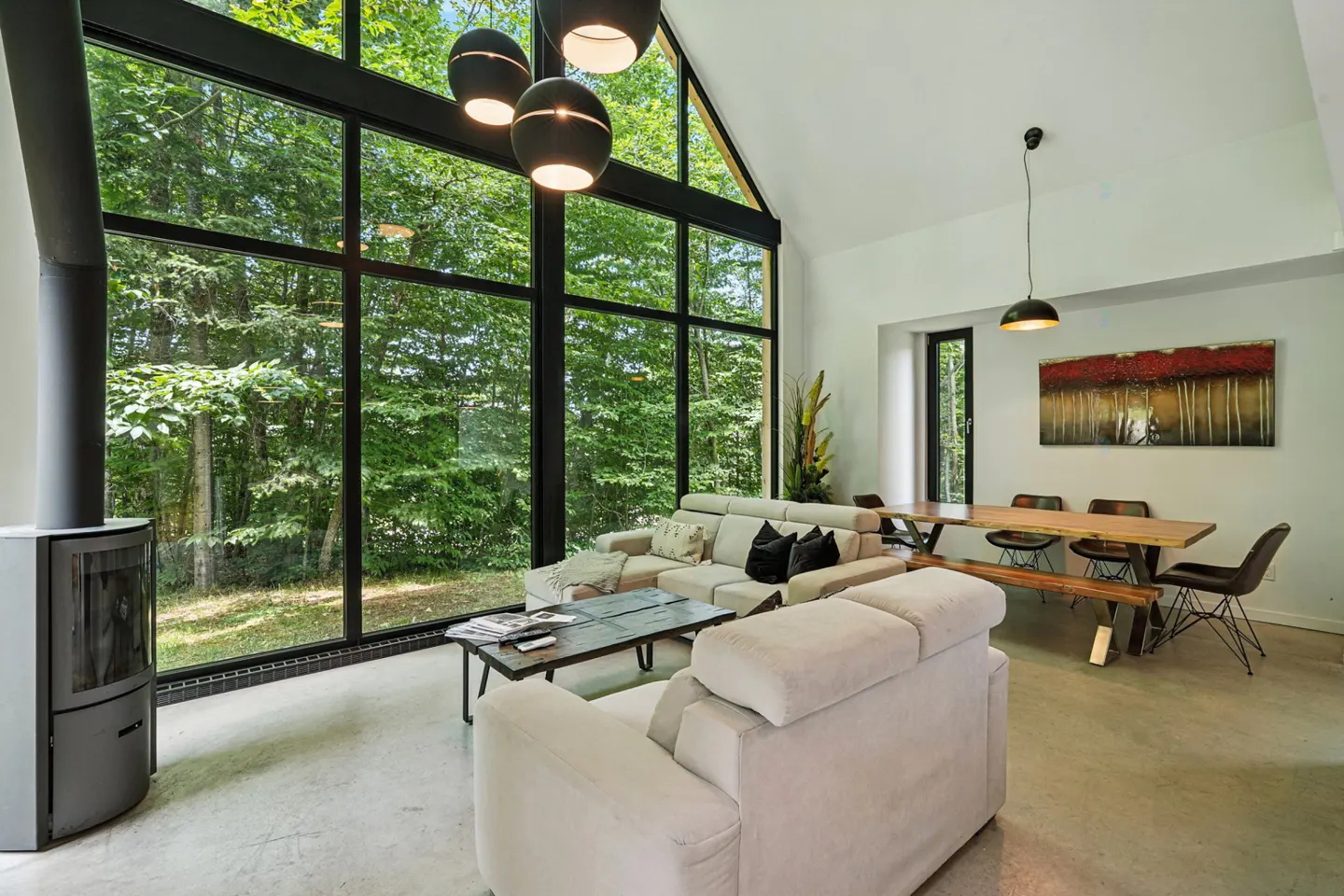 Bright living room with a large window showing a forest. A gray fireplace, beige sofa, and wooden dining table furnish the space.