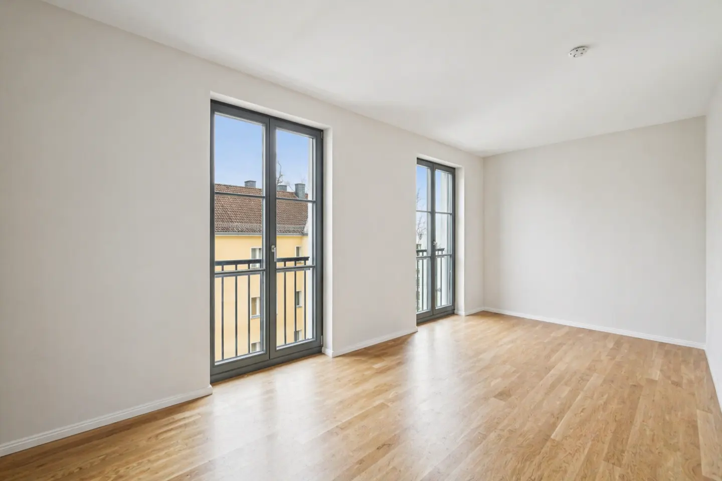 Bright, empty room with hardwood floors, white walls, and two gray-framed windows with balcony views.