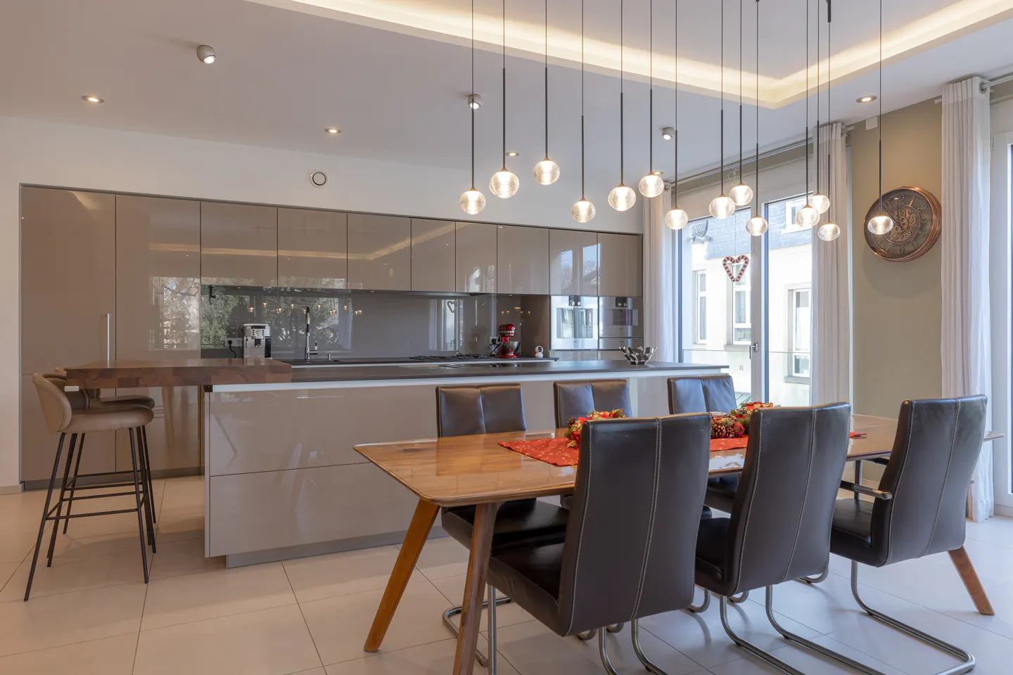 Modern kitchen and dining area with glossy cabinets, wood table, leather chairs, and globe pendant lights.