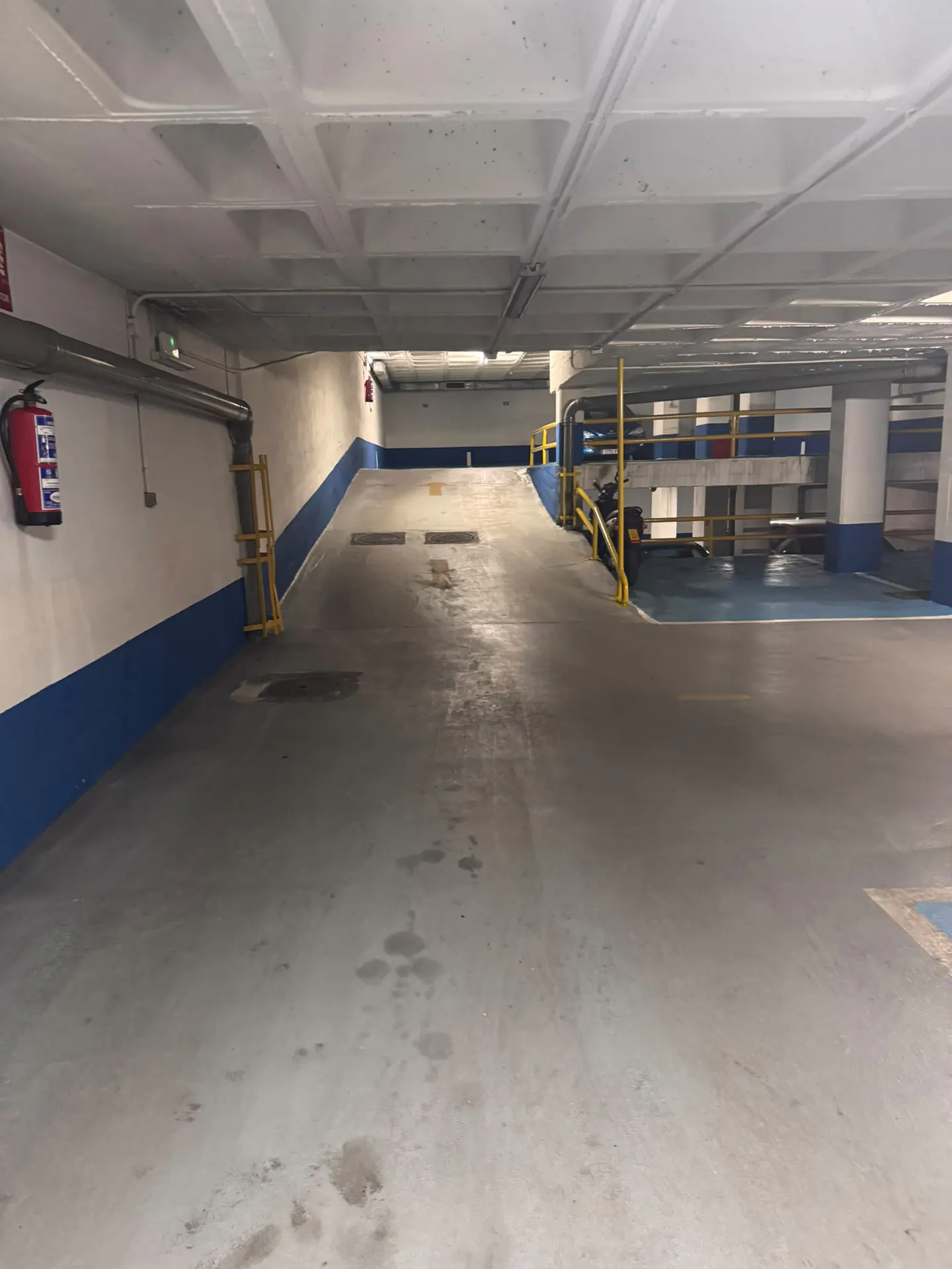 Empty underground parking garage with a concrete ramp, white and blue walls, a red fire extinguisher, and yellow railings.