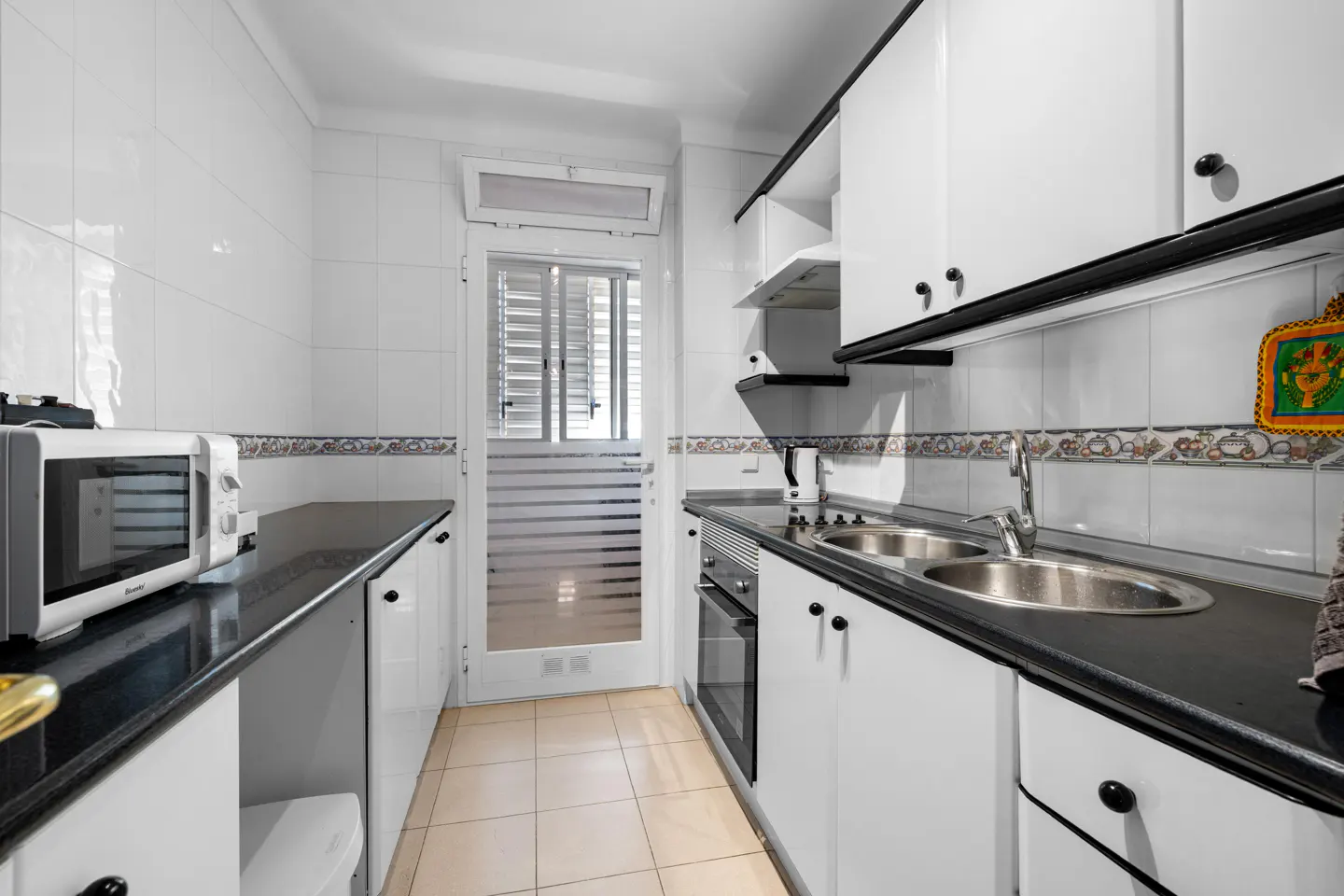 A galley kitchen with white cabinets, black countertops, and tile flooring. A microwave sits on the left counter, and a sink is on the right. A door with blinds is at the end.