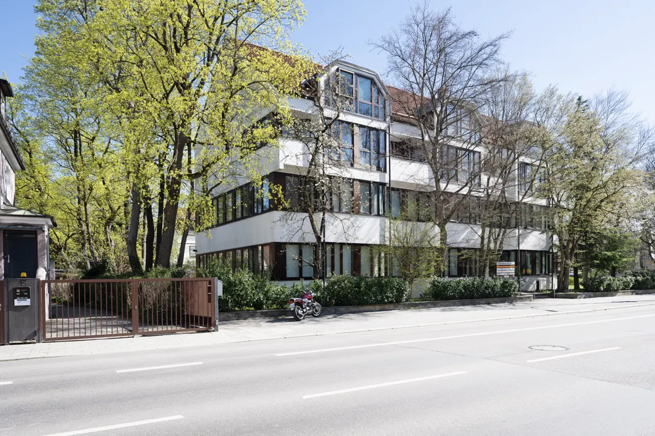A modern, multi-story white building with dark-framed windows, set back from a gray road with trees. A motorcycle is parked near the building.