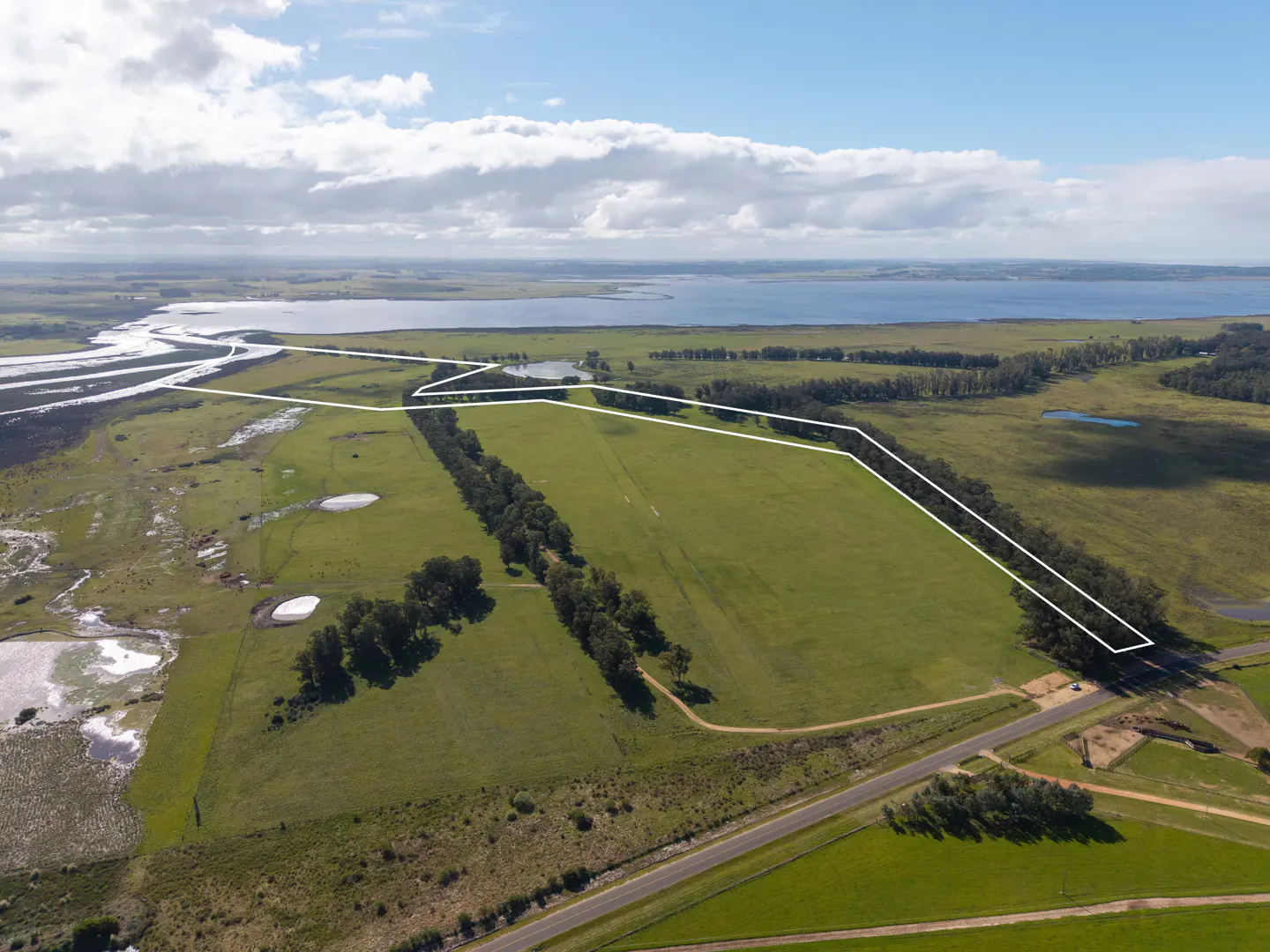 Aerial view of a large green field outlined in white, near a lake and a road under a blue sky.
