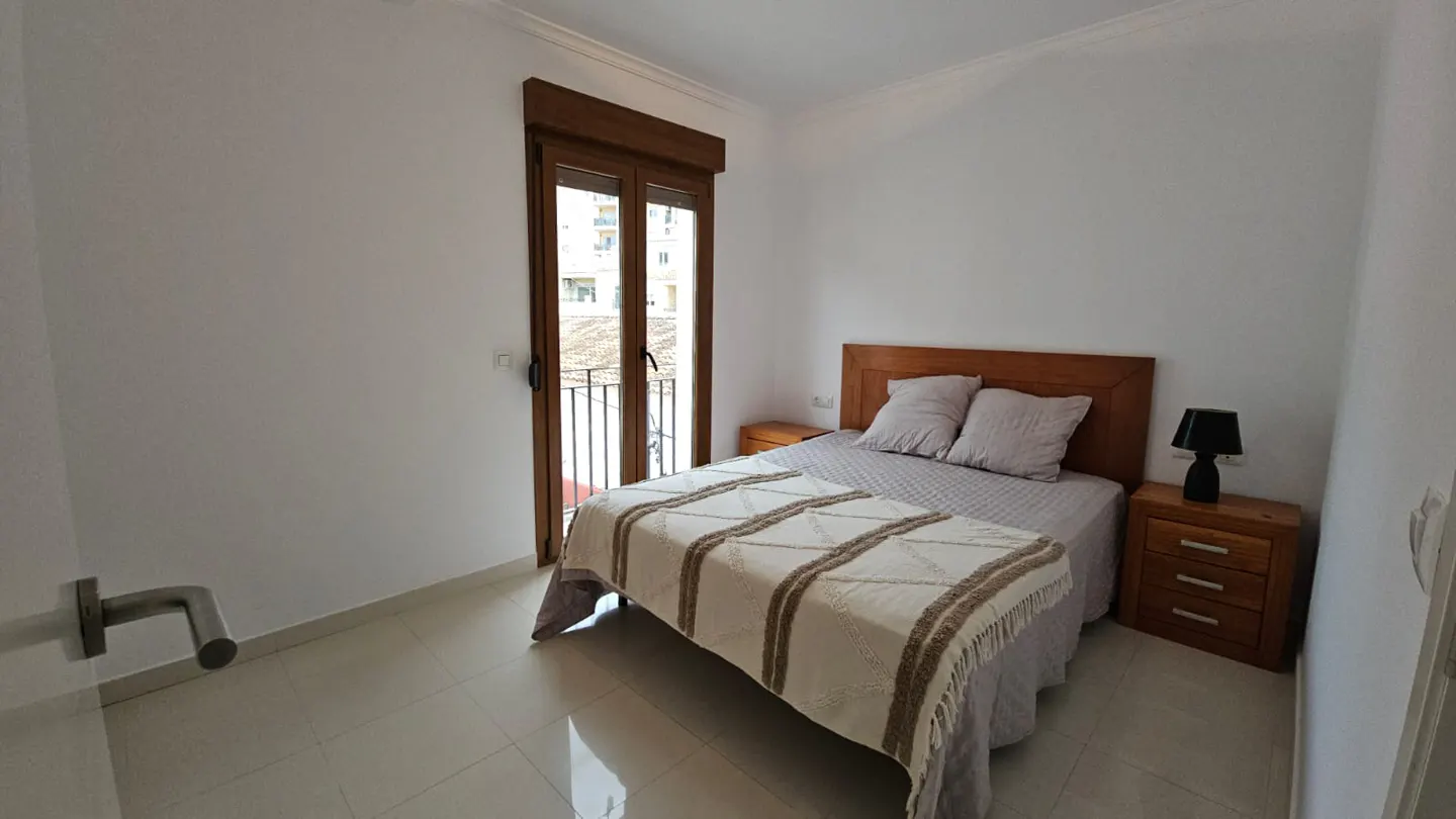 A bright bedroom with white walls, a wooden bed frame, and a balcony door with a view.