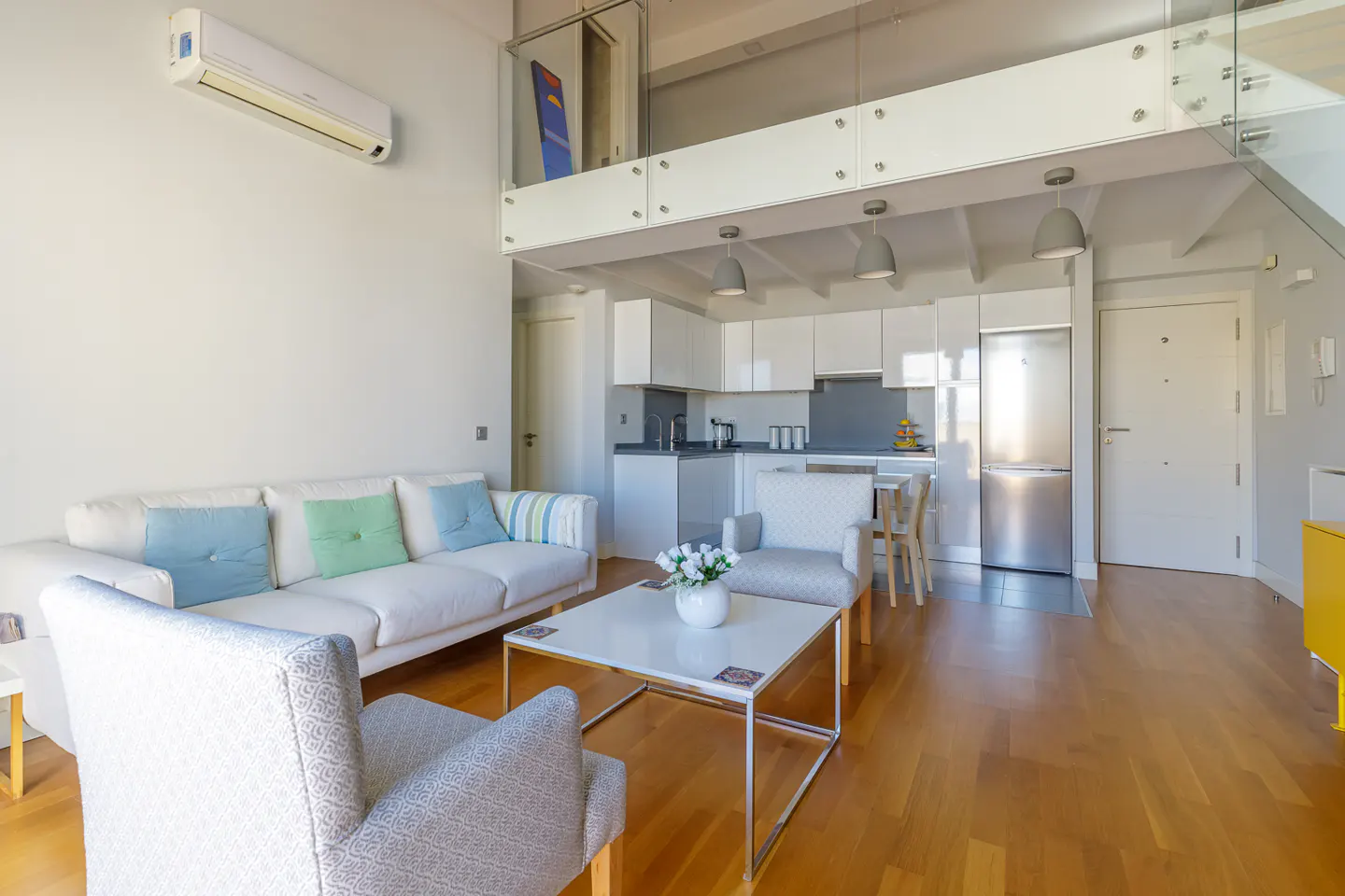 Bright, modern apartment interior with white sofa, patterned armchairs, and a white kitchen under a loft area. Hardwood floors add warmth.