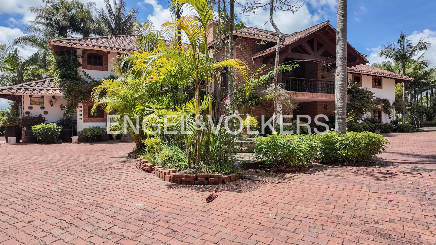 Exterior of a two-story house with a red brick driveway and lush green landscaping. The house has a red tile roof and white walls with brick accents.