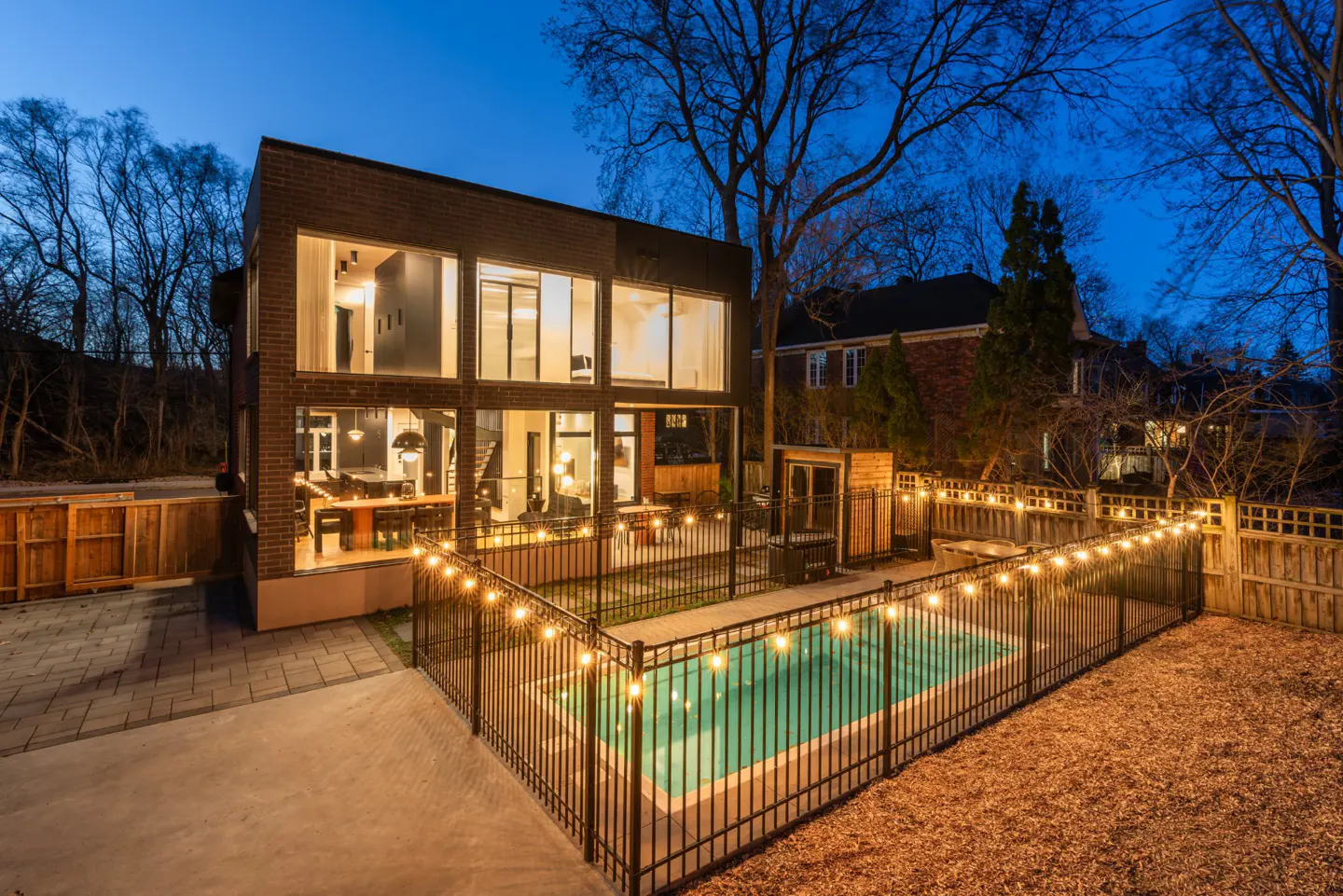 Modern brick house with large windows overlooking a pool, surrounded by string lights at dusk.