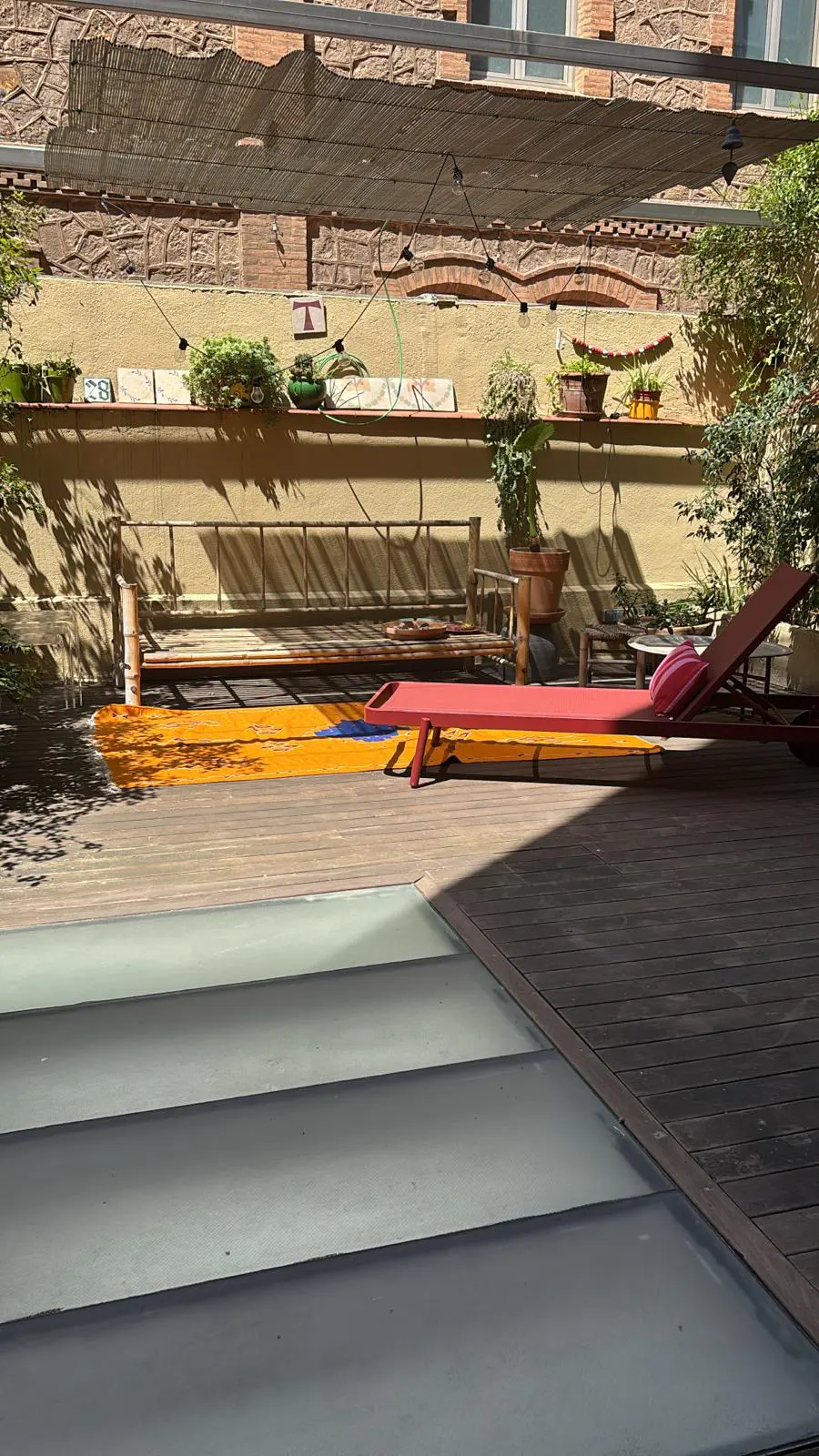 Outdoor patio with wooden deck, bench, lounge chair, and plants against a brick wall under a bamboo shade.