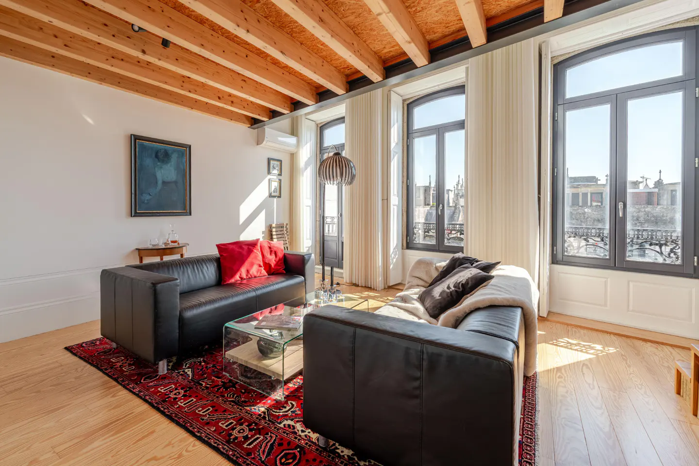 Bright living room with wood floors, exposed beam ceiling, and large windows. Two black leather sofas face each other on a red patterned rug.