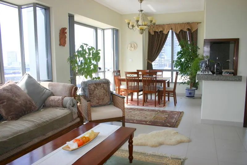 A bright living room with a sofa, chair, dining table, and large windows. A chandelier hangs above the dining area.