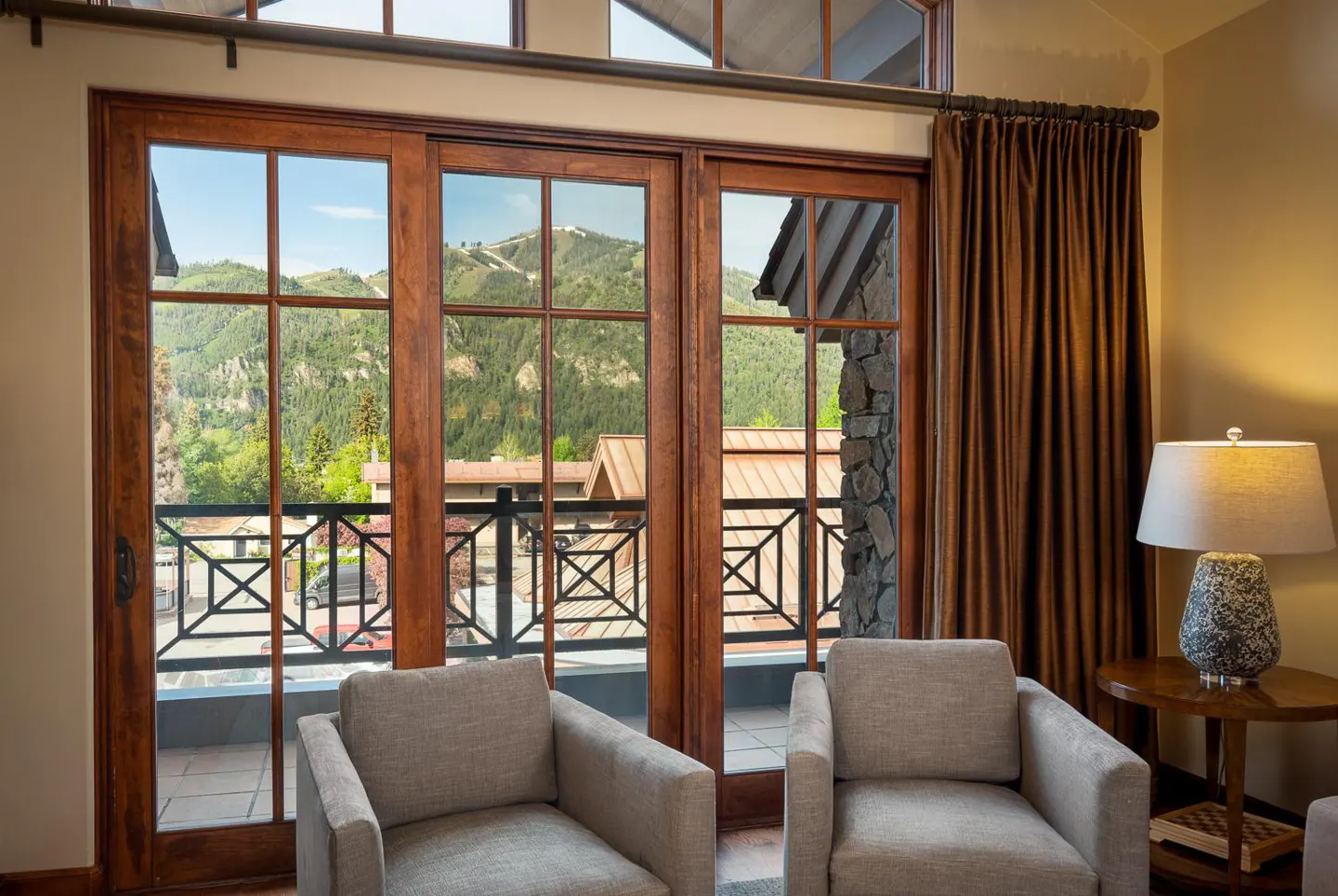 Living room with two armchairs facing a wood-framed sliding glass door with mountain views. Brown curtains and a lamp on a side table.