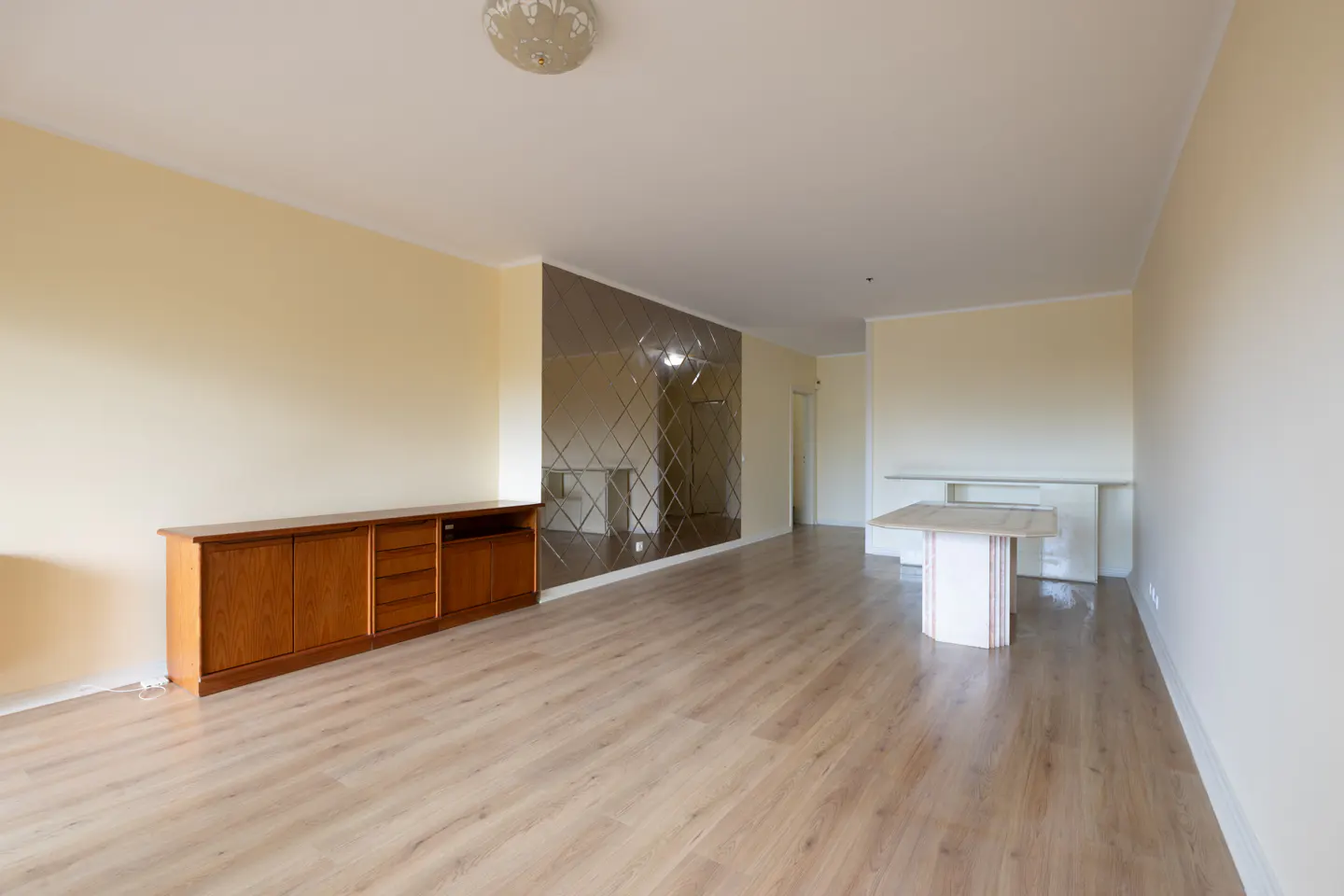 An empty living room with light wood floors, yellow walls, and a brown wooden cabinet. A mirrored wall and a white table are also visible.