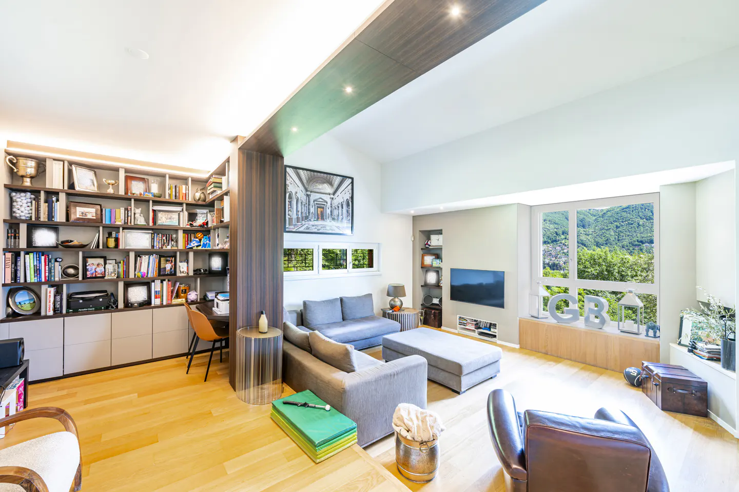 Bright living room with hardwood floors, gray sofas, and a large window with a mountain view. A bookcase filled with books and trophies is on the left.