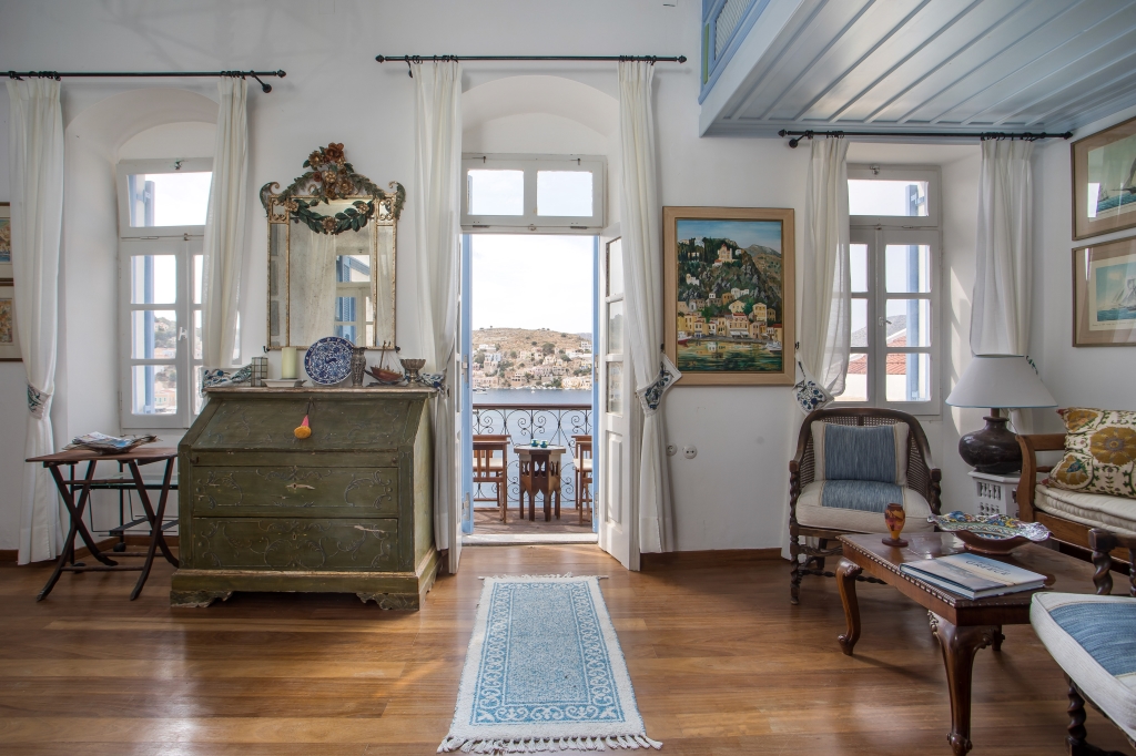 Living room with wooden floors, antique green desk, and open balcony doors revealing a scenic coastal view. [AI-generated text]