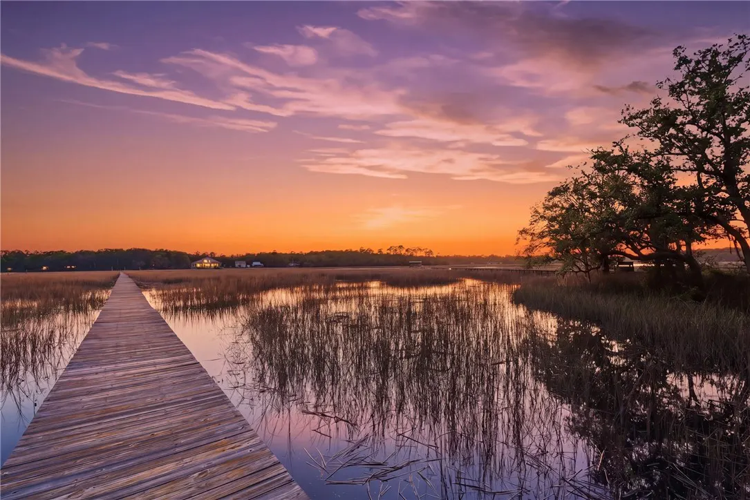 A wooden dock stretches over marshland reflecting a vibrant orange and purple sunset sky. Trees line the right side of the image.