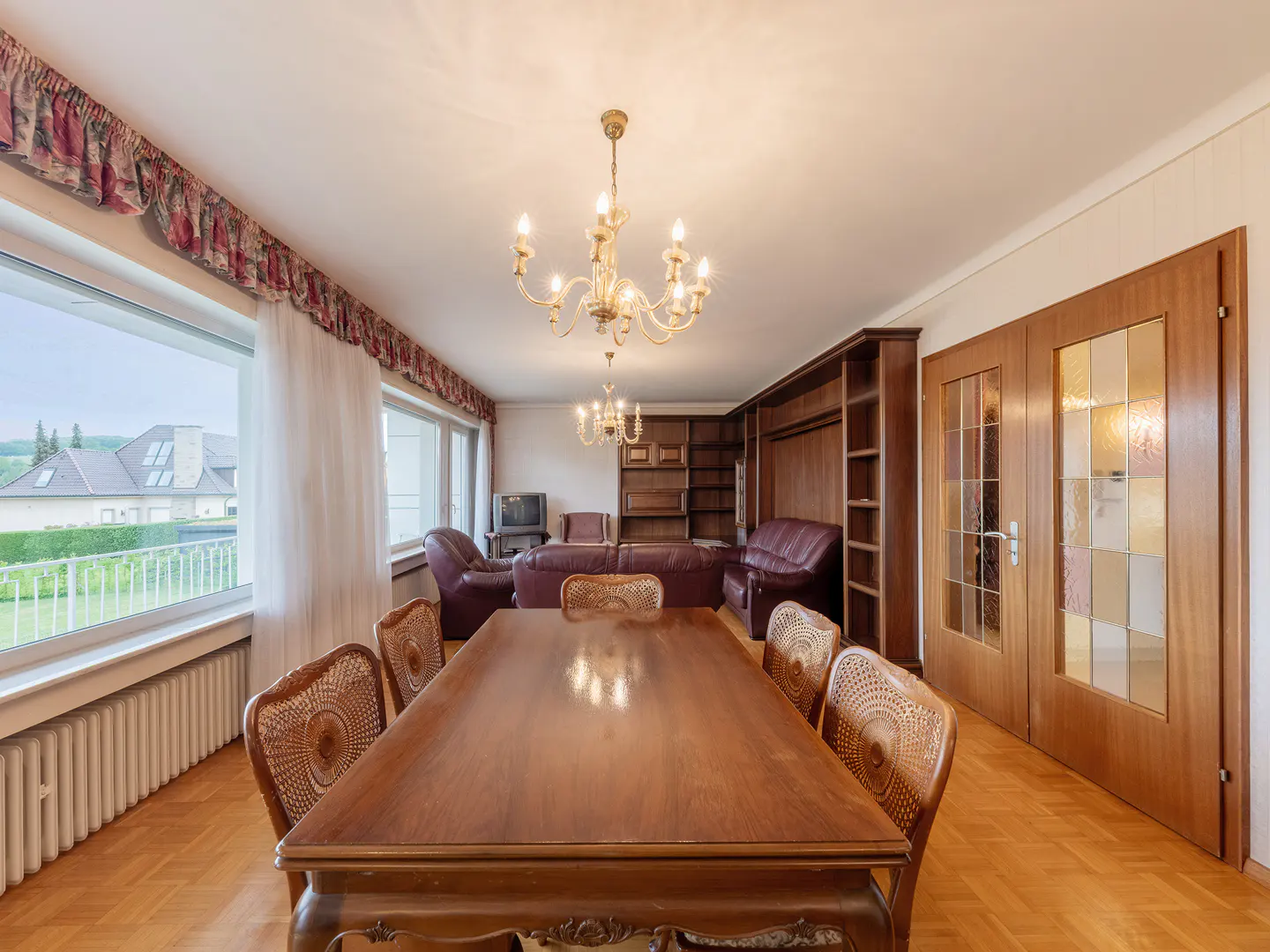 A dining room with a large wooden table and chairs, a chandelier, and a view of the outside.