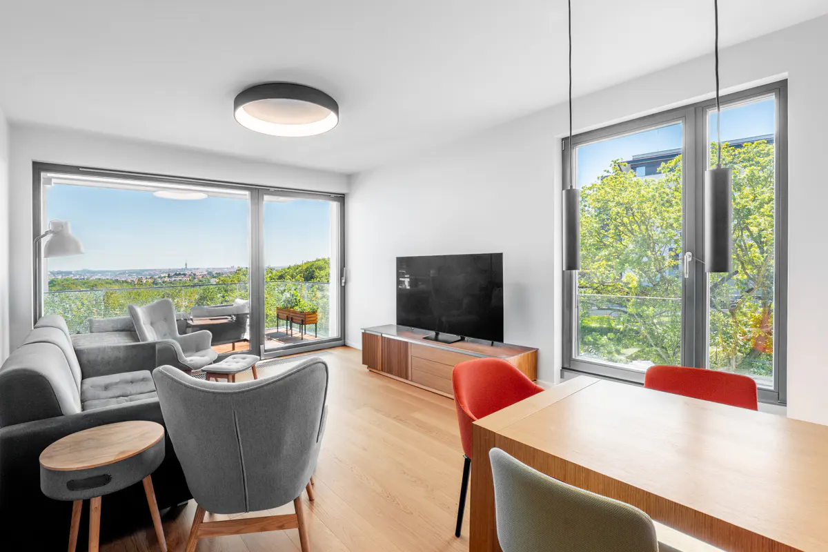 Modern living room with grey sofa, armchairs, wooden dining table with red chairs, and large windows overlooking greenery.