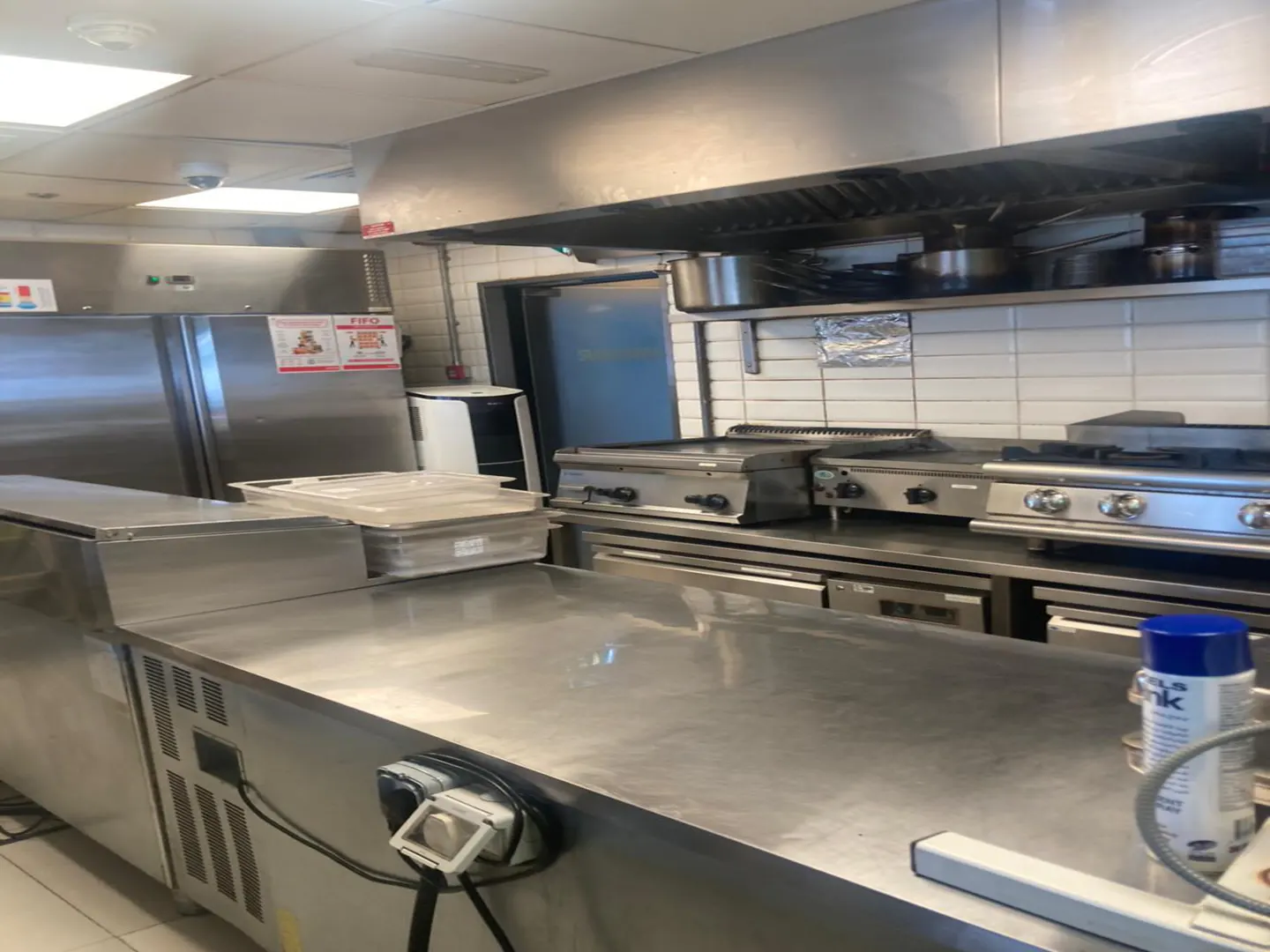 Commercial kitchen with stainless steel counters, industrial appliances, a large refrigerator, and white tiled walls.