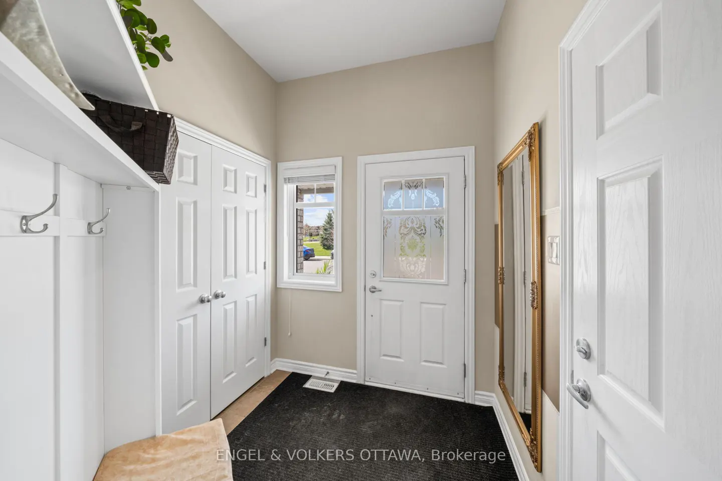 A bright foyer with white walls, a white front door with glass, and a golden framed mirror. A white closet and a bench with hooks are on the left.