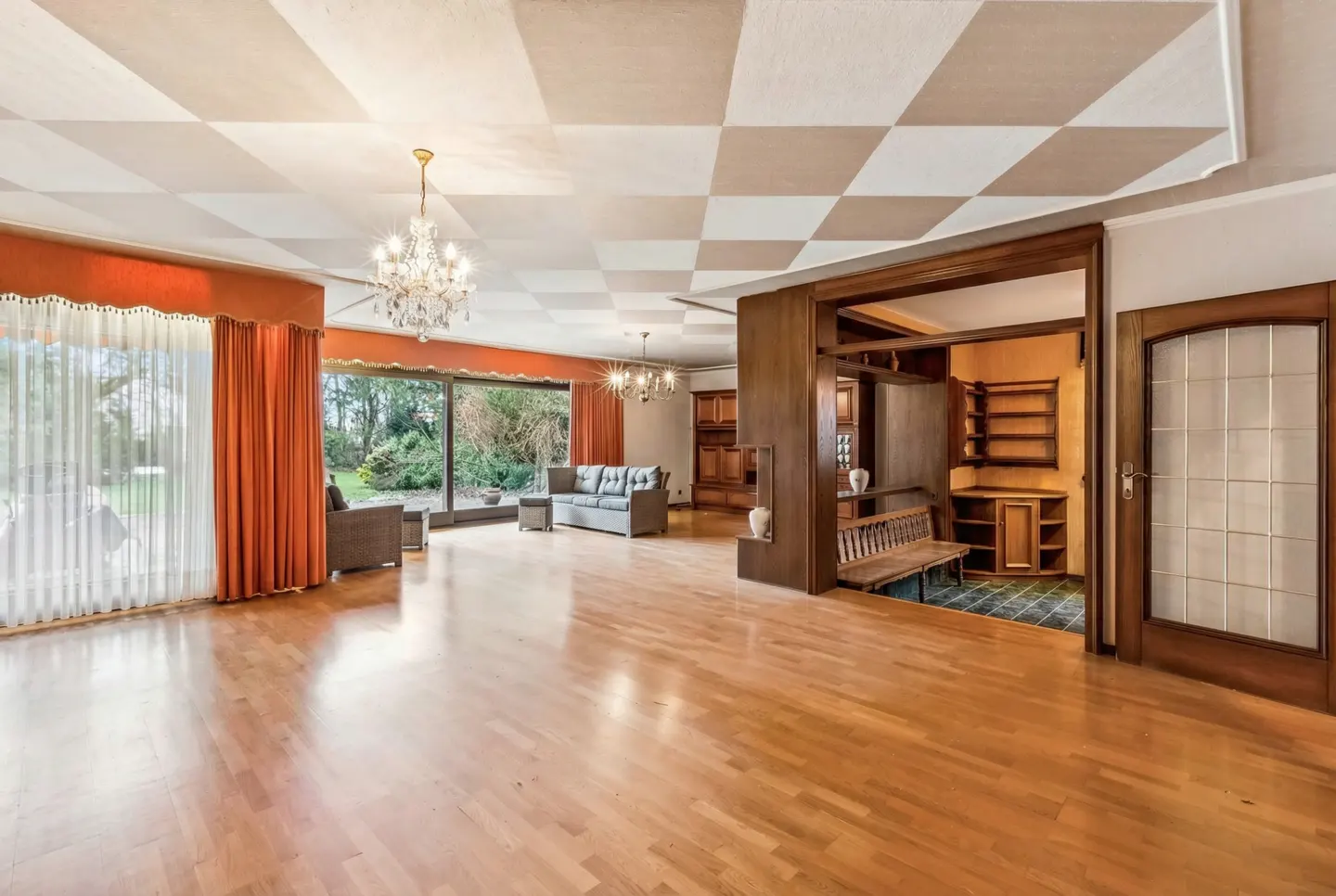 A spacious living room with hardwood floors, orange curtains, and a checkered ceiling. A gray sofa and chairs are visible.