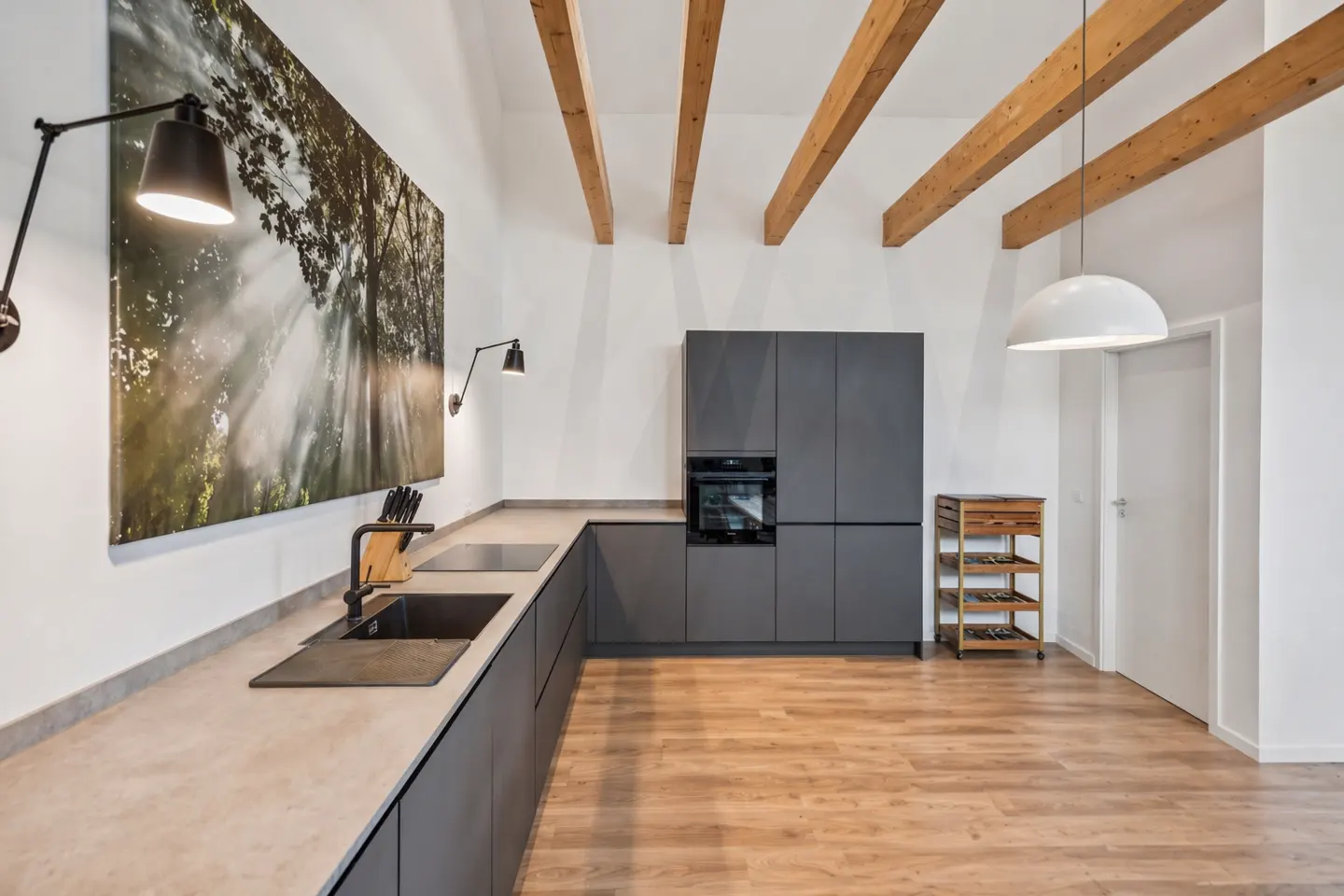Modern kitchen with gray cabinets, wood floors, and exposed beams. A large nature print hangs on the wall with black sconces.