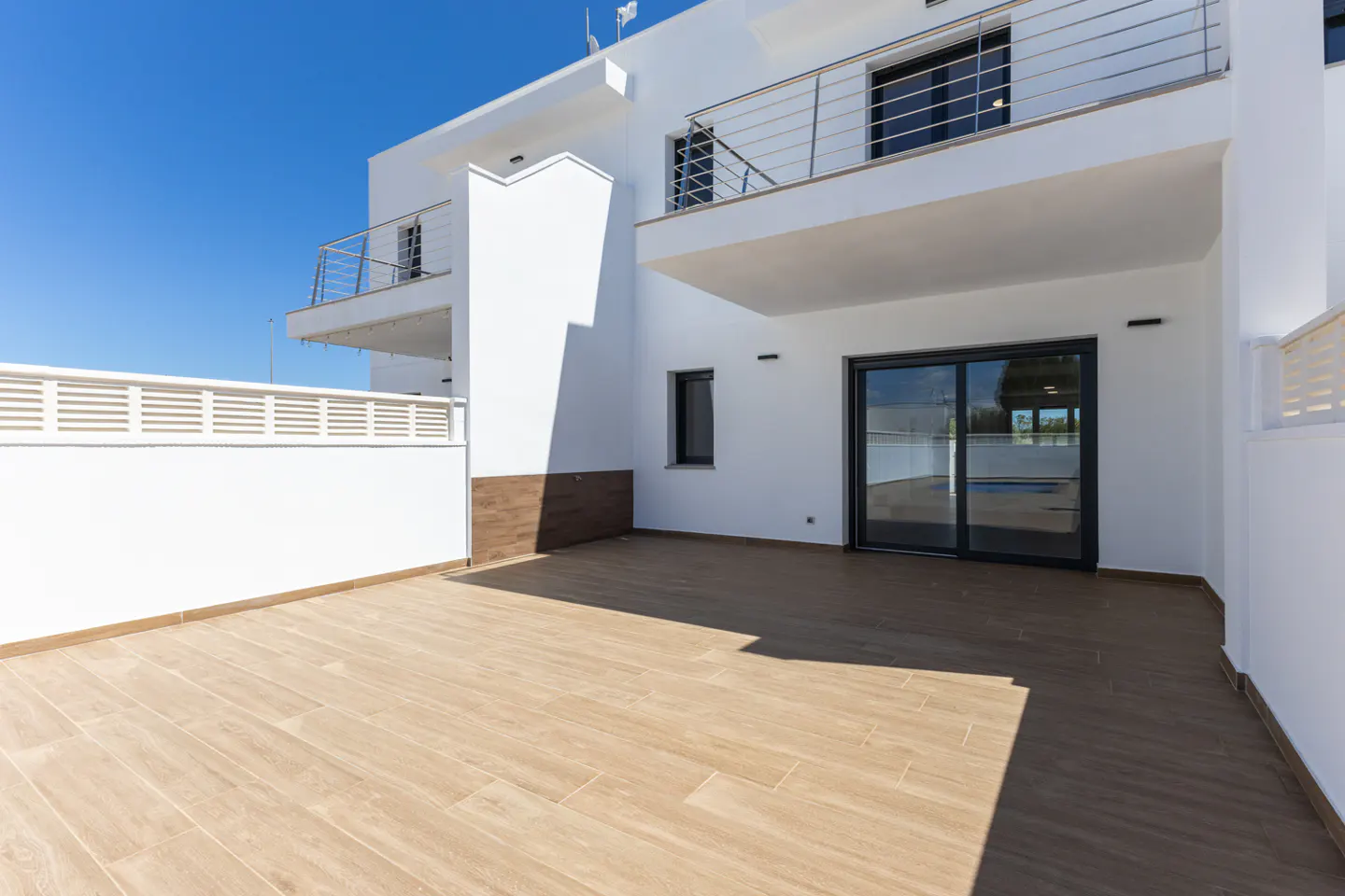 Exterior view of a modern, white two-story house with a wood-tiled patio and sliding glass doors. Balconies with metal railings are visible.