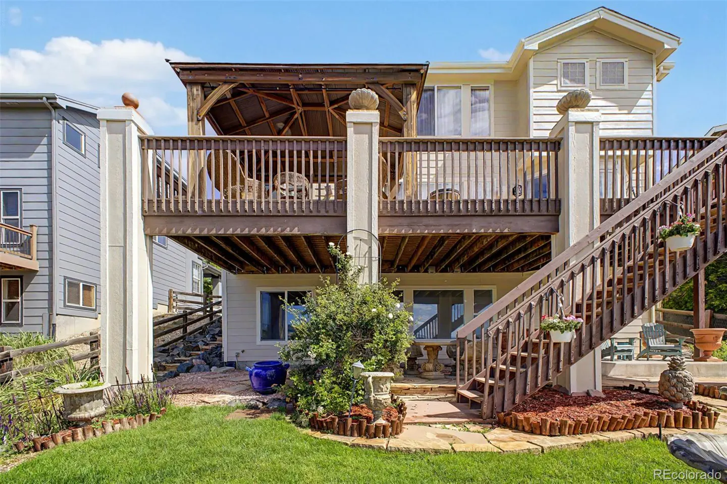 Backyard of a tan house featuring a large wooden deck with a pergola, stairs, and a landscaped patio area with grass.