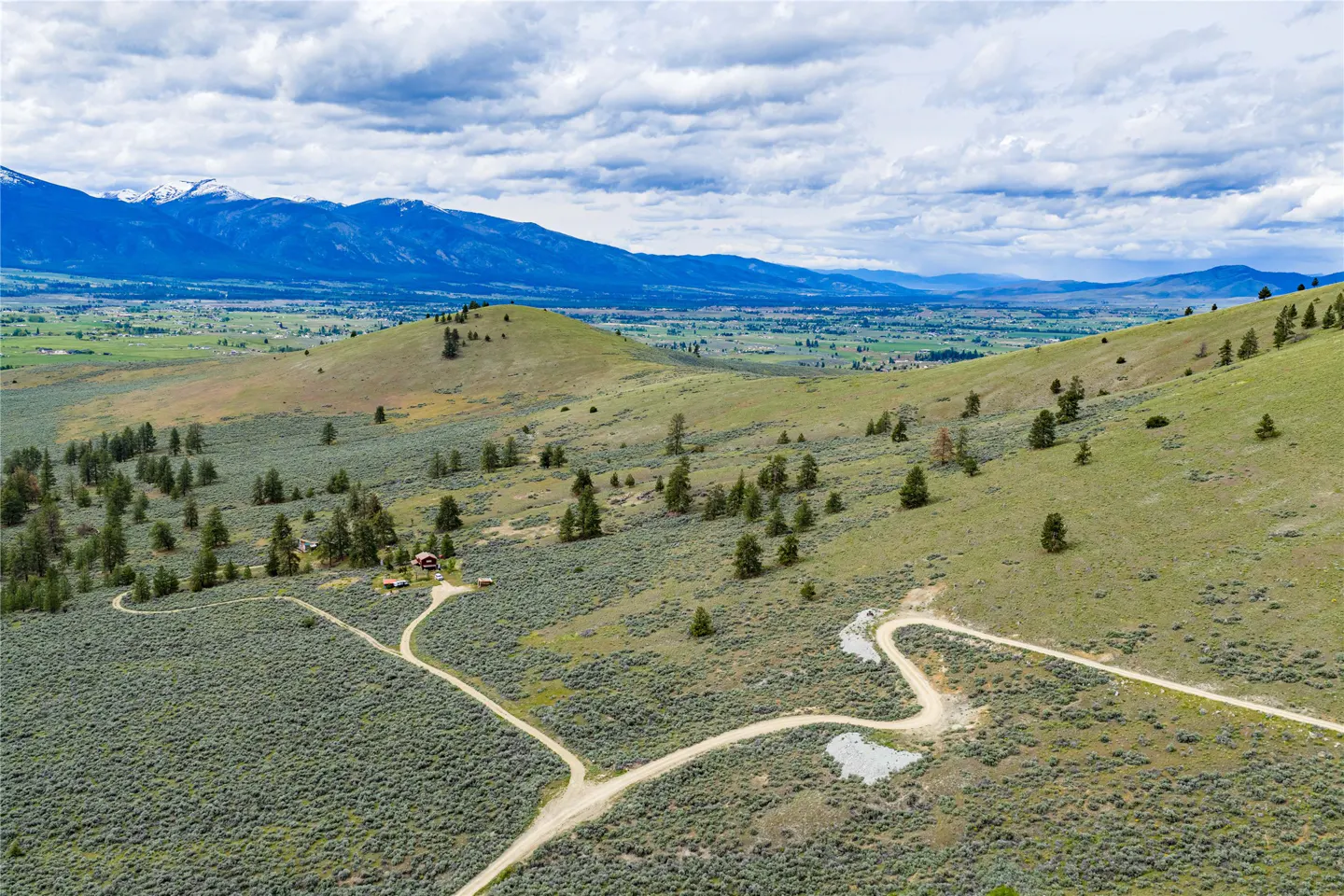Aerial view of a green, hilly landscape with a dirt road leading to a small house, mountains in the distance.