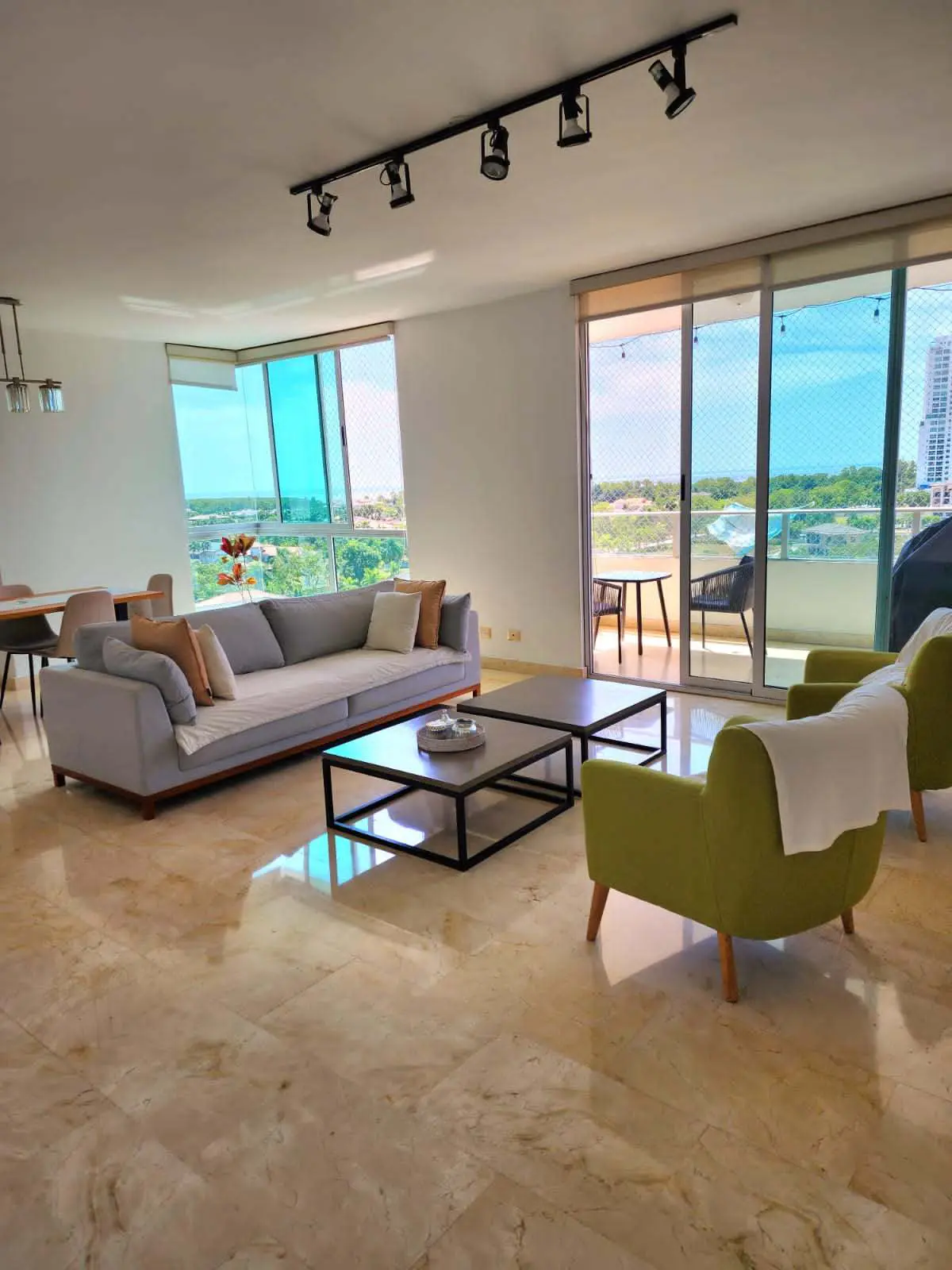 Bright living room with a gray sofa, green chairs, and two black coffee tables. A balcony with furniture overlooks a scenic view.
