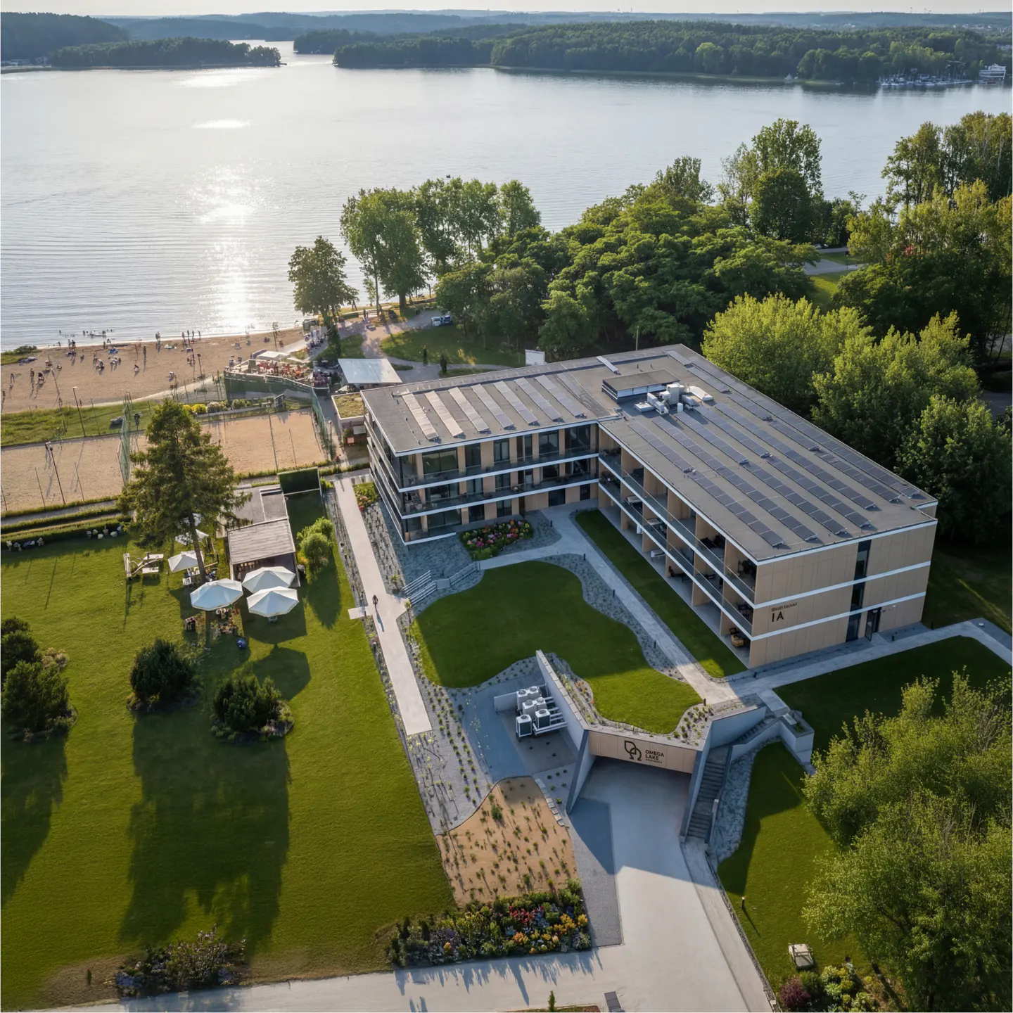 Aerial view of a modern multi-story building with solar panels, set on a lush green lawn beside a sandy beach and lake.