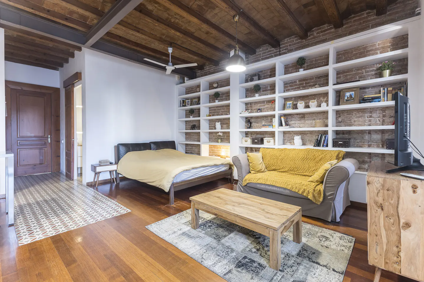 Bedroom with wooden floors, exposed brick wall with white shelves, bed, sofa, and wooden coffee table.